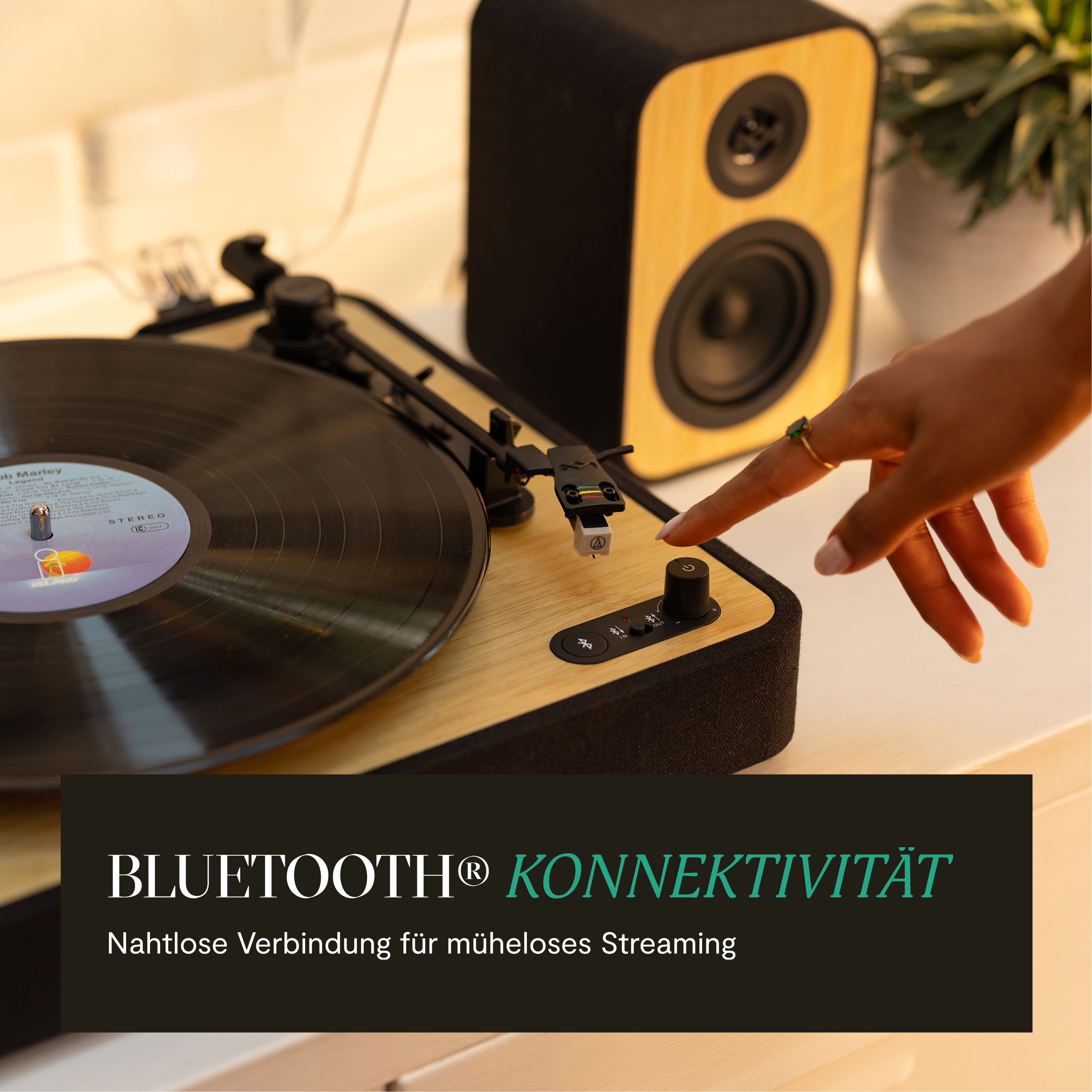 Eine Hand berührt einen Plattenspieler. Im Hintergrund befinden sich ein Lautsprecher und eine Schallplatte. Text: 'Bluetooth Konnektivität'.