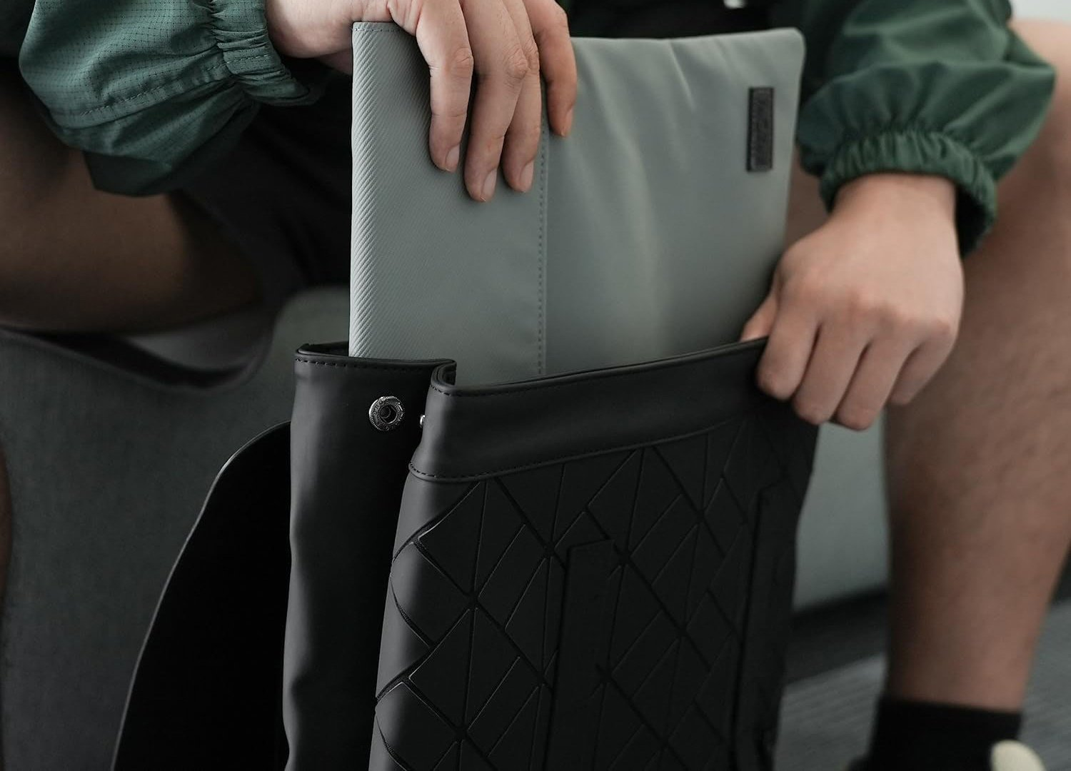 Una persona inserisce un laptop grigio in uno zaino nero. Lo zaino ha un motivo geometrico. La persona indossa una giacca verde.
