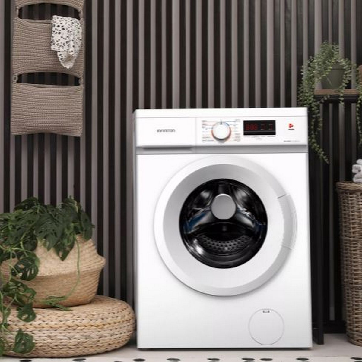 Lavadora blanca en una habitación. Plantas, cestas y una pared a rayas en el fondo. Un pequeño botón redondo está en la máquina.
