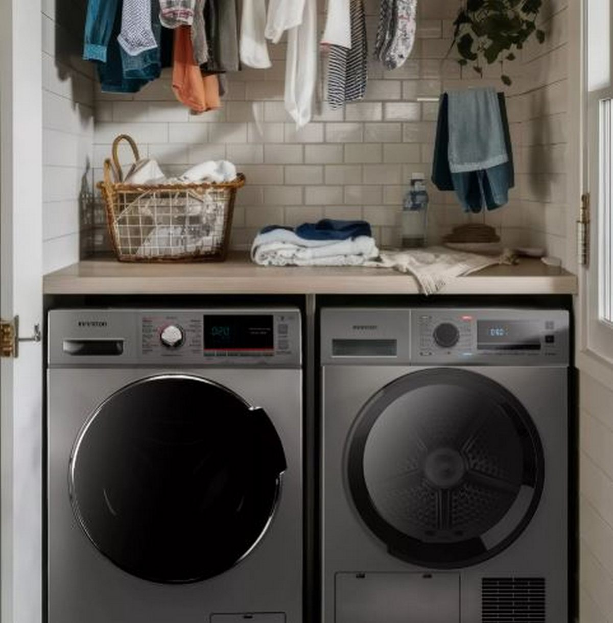 Cuarto de lavado con dos lavadoras plateadas. Ropa colgada arriba, una cesta de artículos sobre la encimera, con ropa doblada.