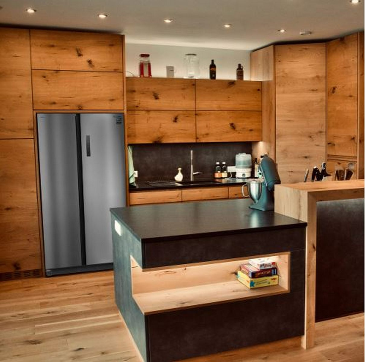 Cocina moderna con armarios de madera, nevera de acero inoxidable, isla negra y batidora. Libros en estantería.