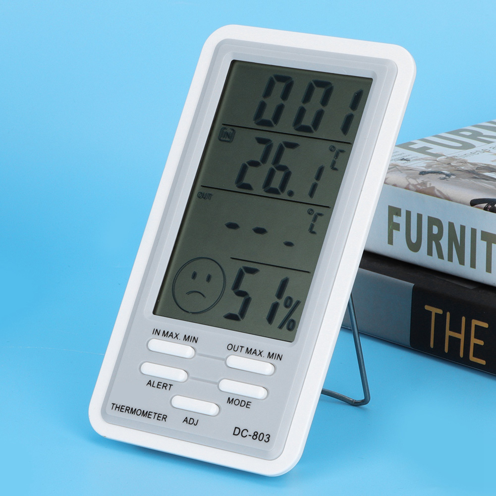 Ein digitales Thermometer zeigt Temperatur und Luftfeuchtigkeit an. Ein trauriges Gesichtssymbol ist sichtbar. Bücher sind im Hintergrund.