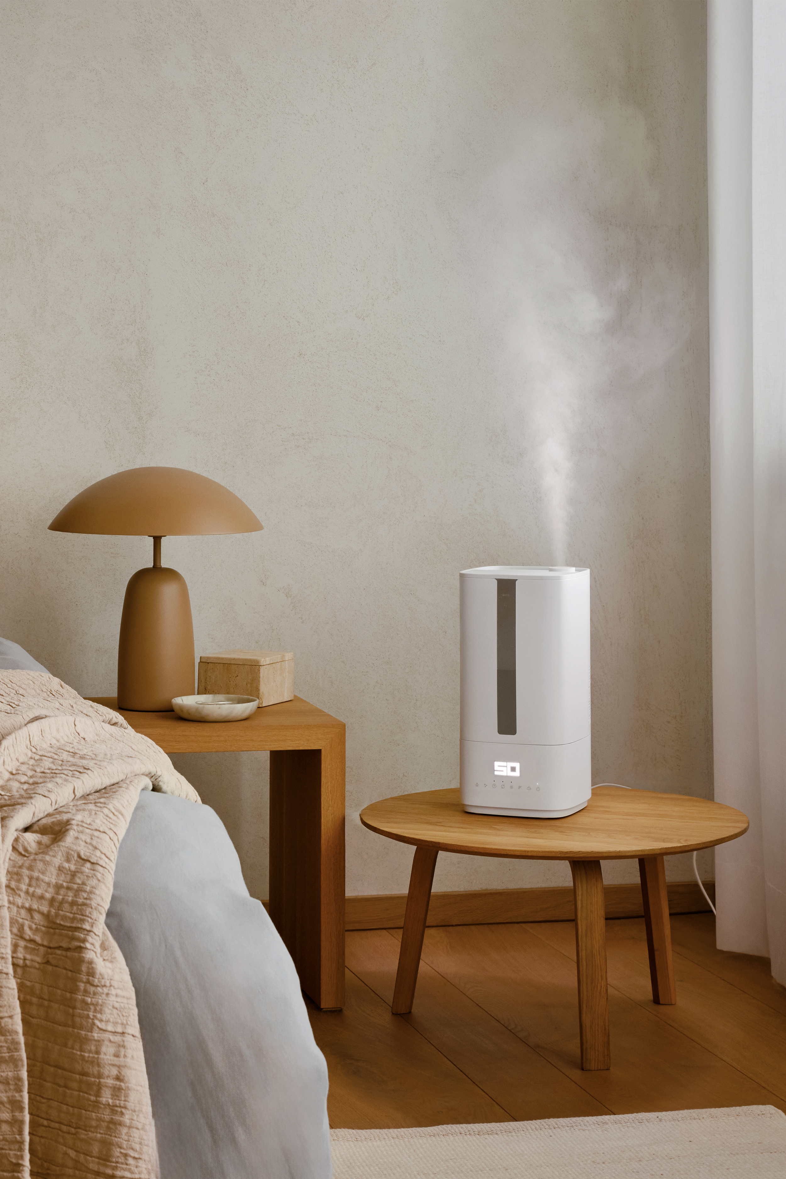 A bedroom scene. A lamp and a box sit on a wooden table near a bed. A humidifier is on another table.
