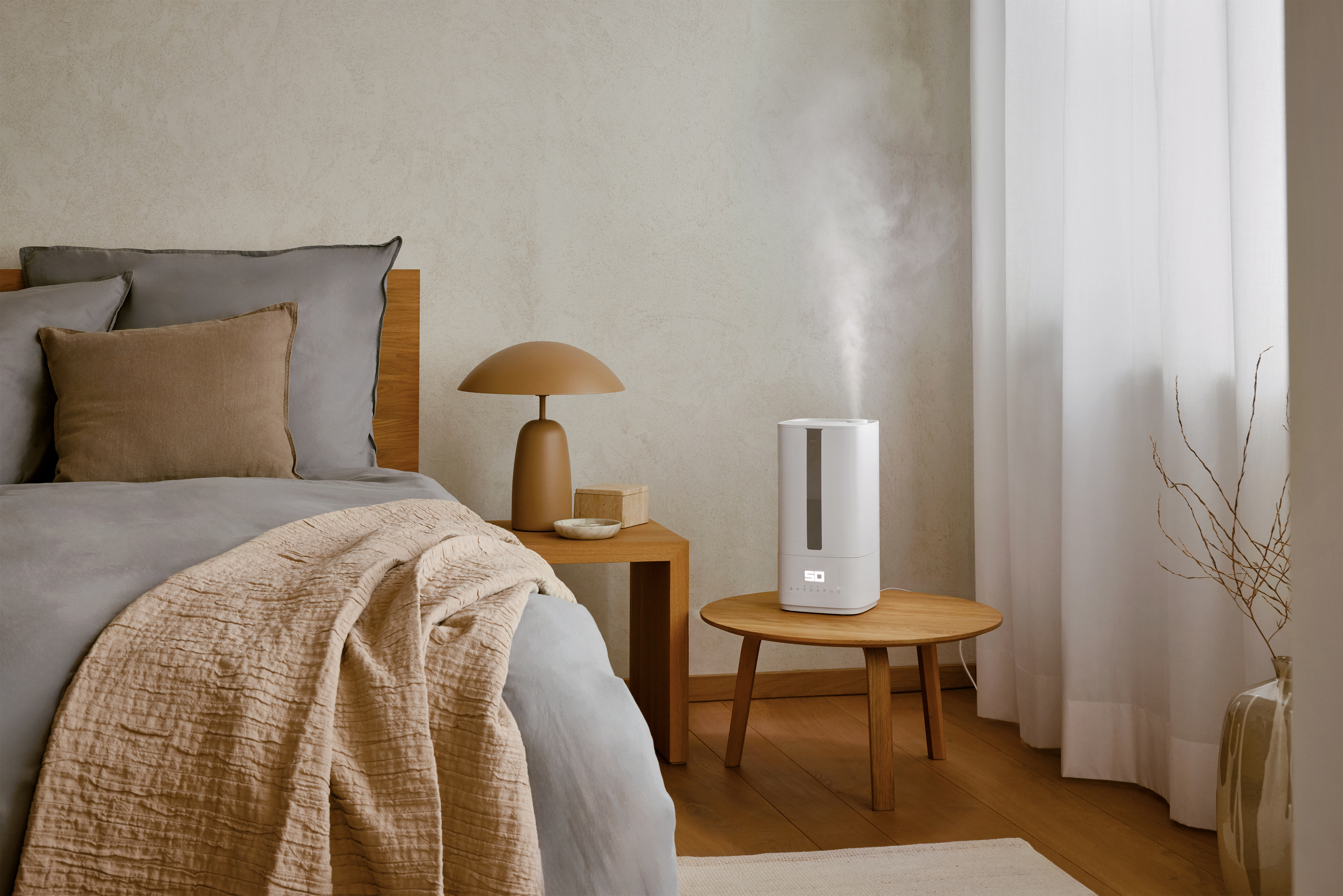A bedroom scene with a bed, table lamp, and humidifier. Neutral tones dominate the space, creating a calm atmosphere.