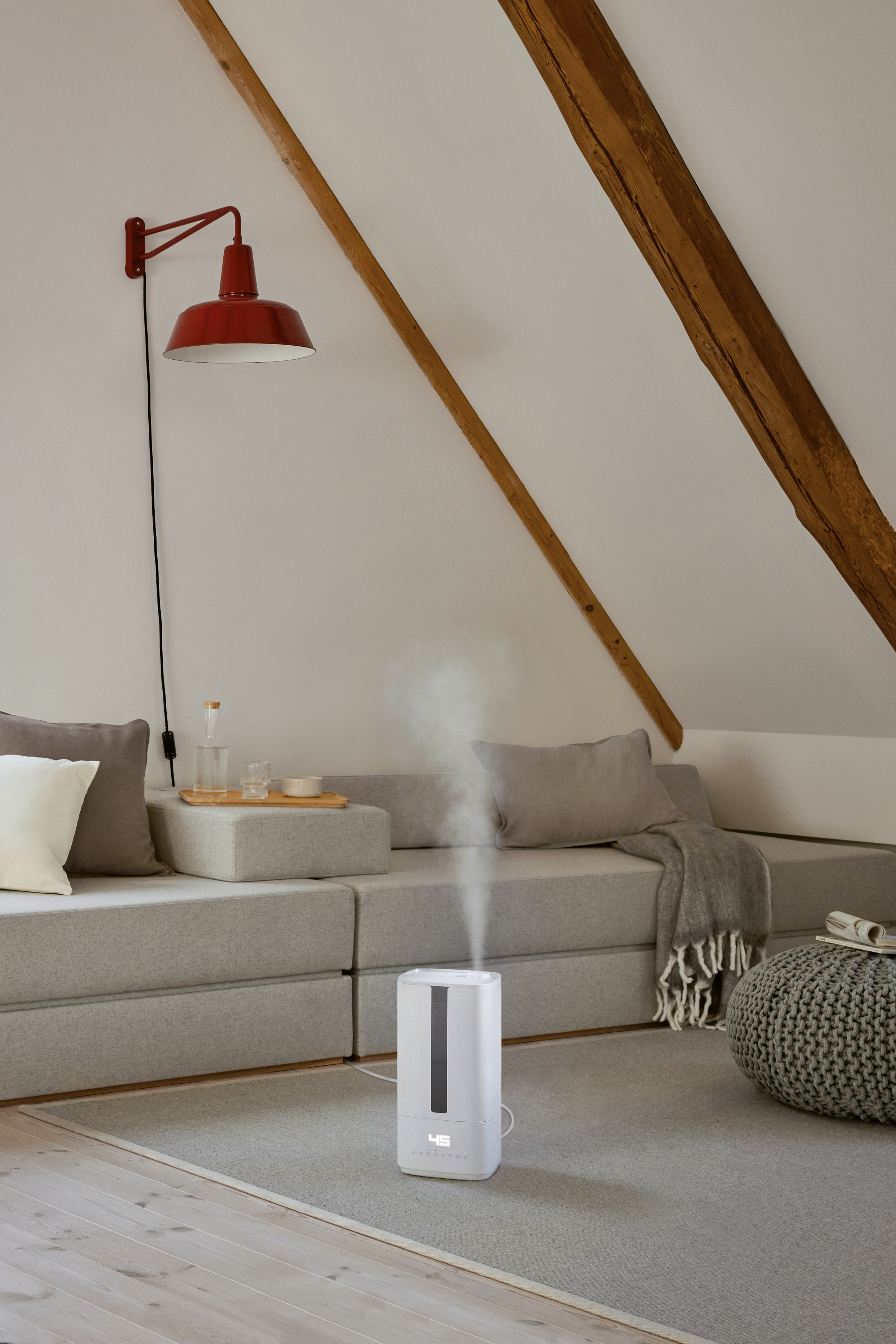 A white humidifier emits steam in a room. A red lamp hangs above a gray sofa with pillows and a blanket.
