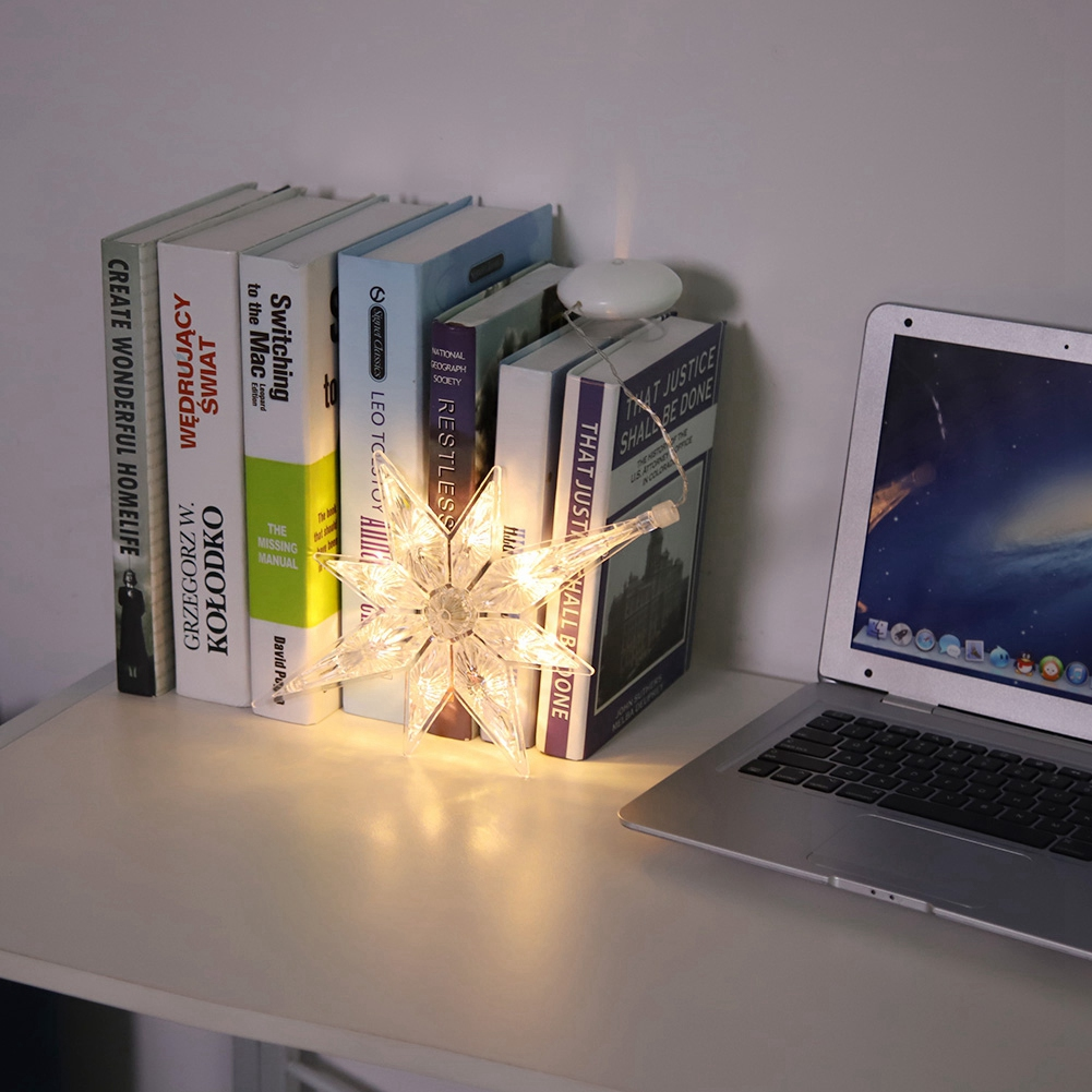 Eine beleuchtete Sternendekoration steht auf einem Schreibtisch neben Büchern und einem Laptop. Die Bücher stehen aufrecht. Die Szene ist gut beleuchtet.