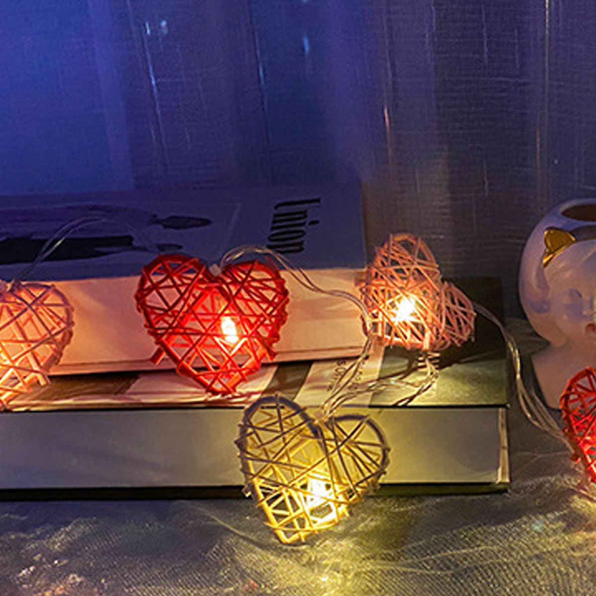 Herzförmige Lichterkette, rot und gelb, auf Büchern. Sanftes Leuchten. Hintergrund: Vorhang und ein kleines Keramikobjekt.