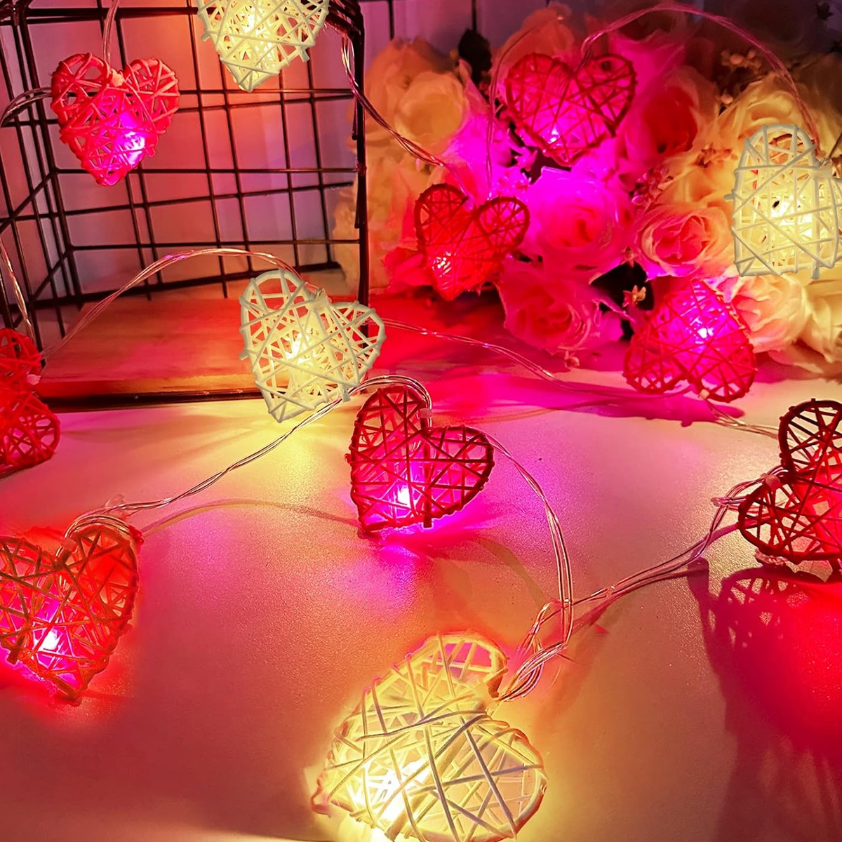 Lichterketten mit roten und weißen Herzformen. Im Hintergrund ein Blumenstrauß. Die Szene ist mit rosa Licht beleuchtet.