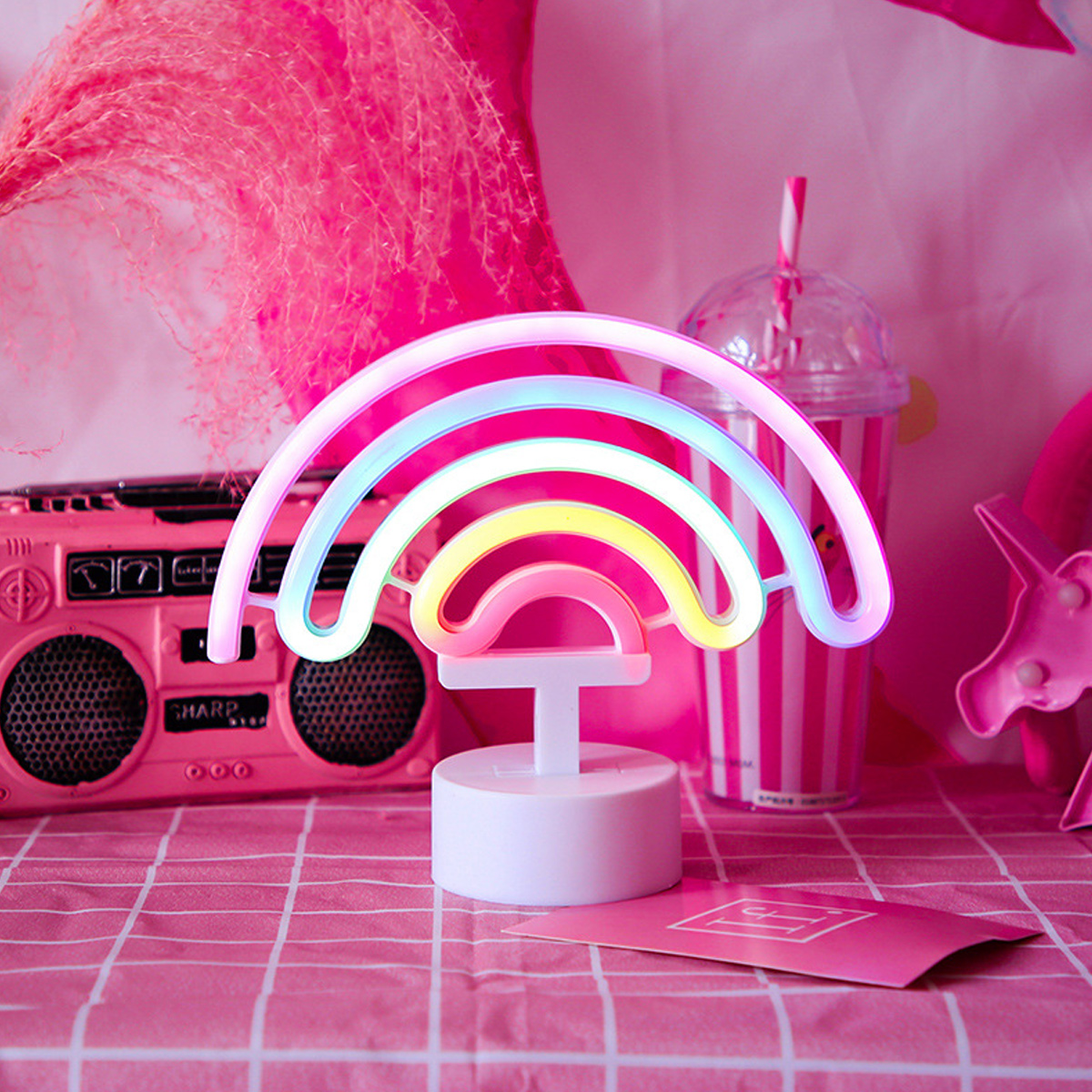 Ein rosa Neon-Regenbogenlicht steht auf einem Tisch mit einem rosa Ghettoblaster, einem Getränk und einer rosa Karte. Der Hintergrund ist rosa.