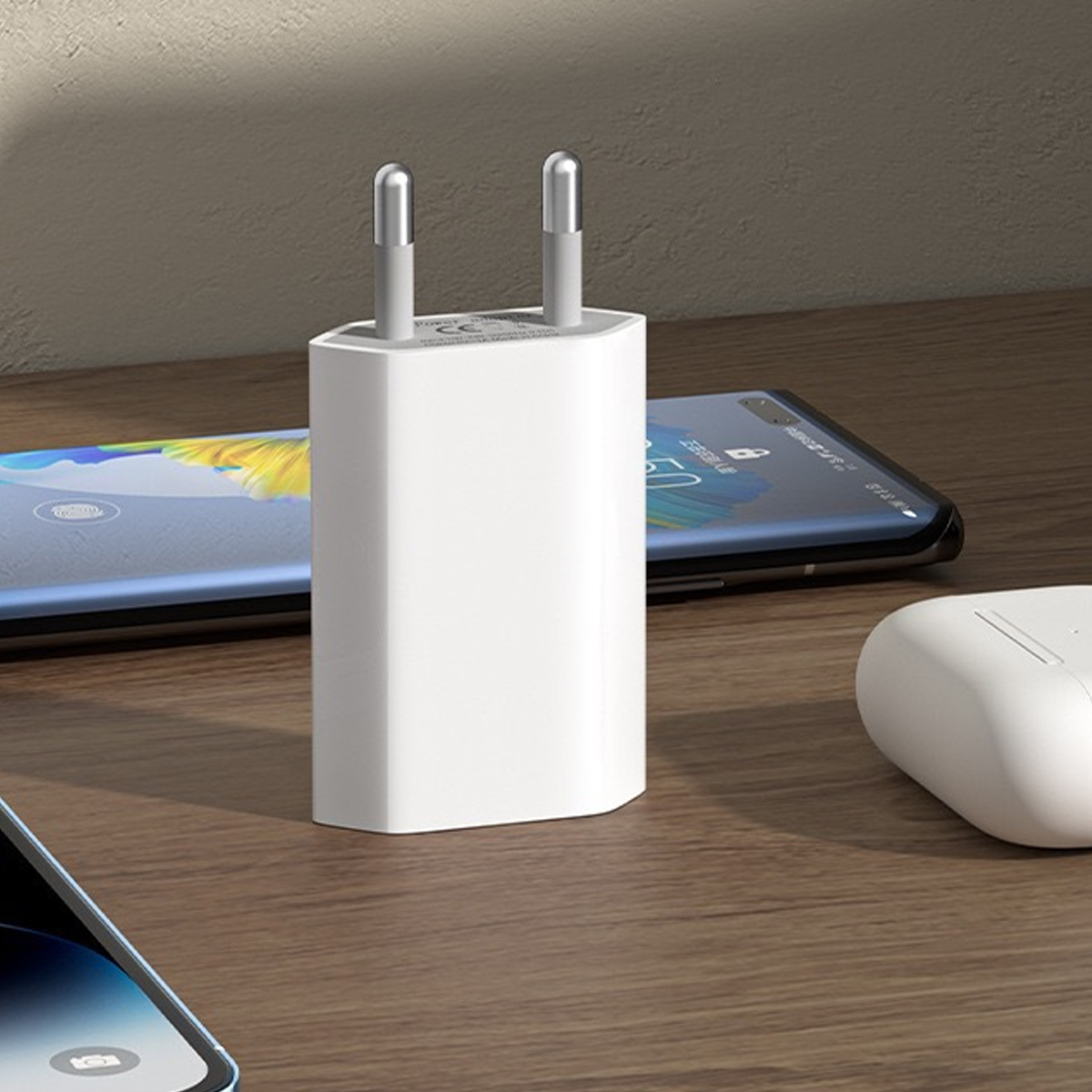Ein weißes Telefonladegerät mit zwei Metallstiften steht auf einer Holzoberfläche. Zwei Smartphones und ein weißes Ohrhörergehäuse sind in der Nähe.