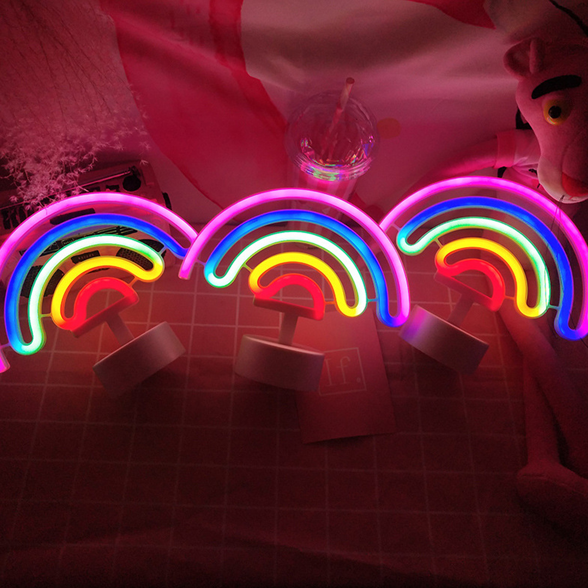 Drei Neon-Regenbogenlichter auf einem Tisch. Pink Panther Stofftier und ein Getränk mit Strohhalm im Hintergrund.
