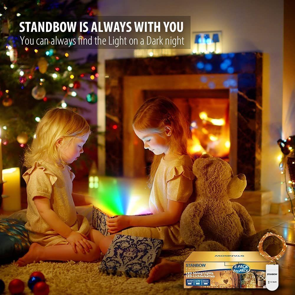 Zwei Mädchen schauen auf eine Kiste, die farbiges Licht aussendet. Teddybär, Kamin und Weihnachtsbeleuchtung im Hintergrund. Produktverpackung sichtbar.