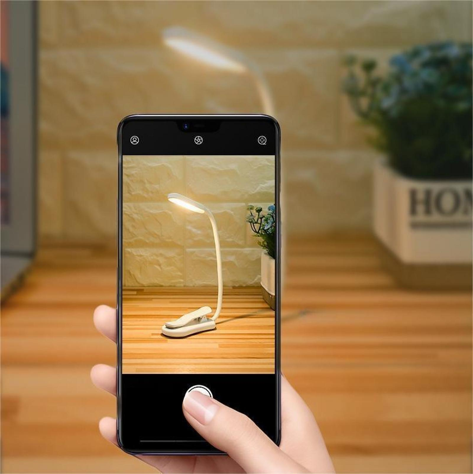 Eine Hand hält ein Telefon, das eine Schreibtischlampe zeigt. Die Lampe ist weiß und hat einen Clip. Hintergrund: eine Wand und eine Pflanze.