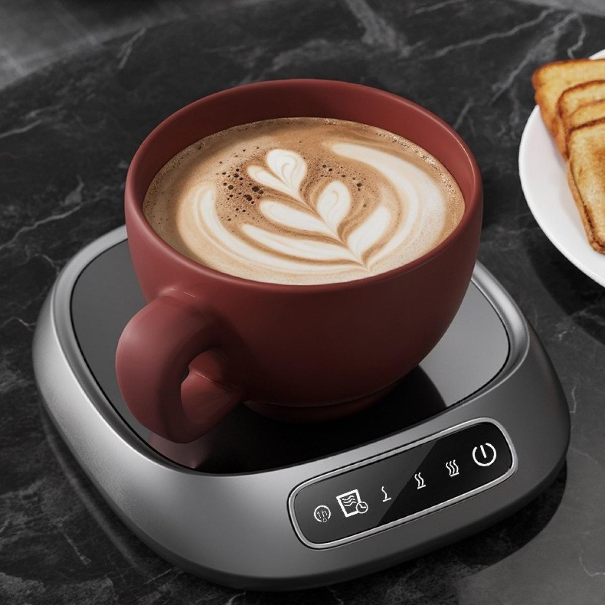 Eine kastanienbraune Kaffeetasse mit Latte Art steht auf einem grauen Wärmer, neben Toast auf einem weißen Teller, alles auf schwarzem Marmor.