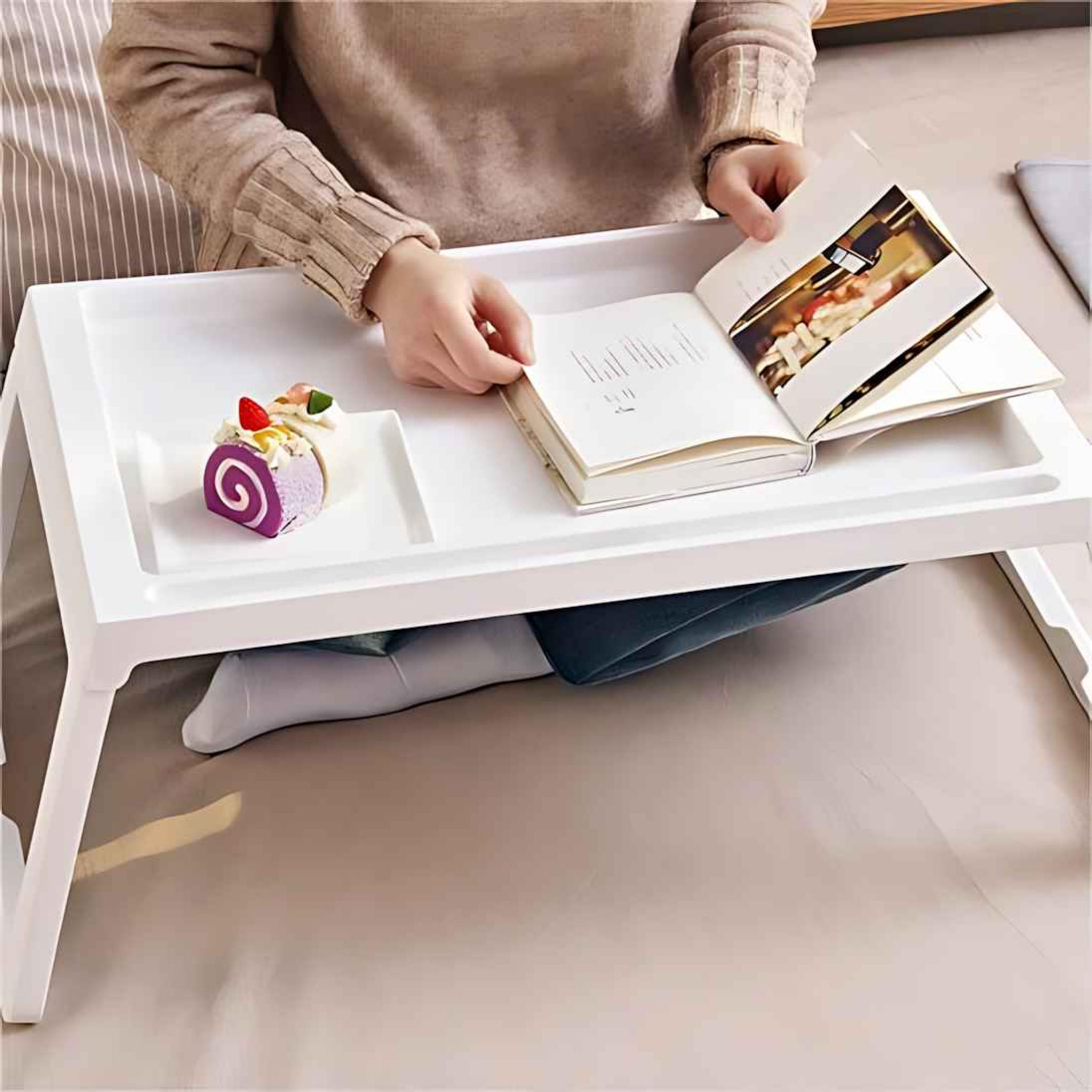 Eine Person liest ein Buch auf einem weißen Tablett mit Essen. Das Tablett steht auf Beinen. Auf dem Tablett liegt ein Kuchen.