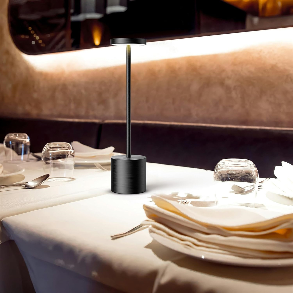 Eine schwarze Lampe steht auf einer weißen Tischdecke in einem Restaurant. Auf anderen Tischen sind Gläser und Besteck.