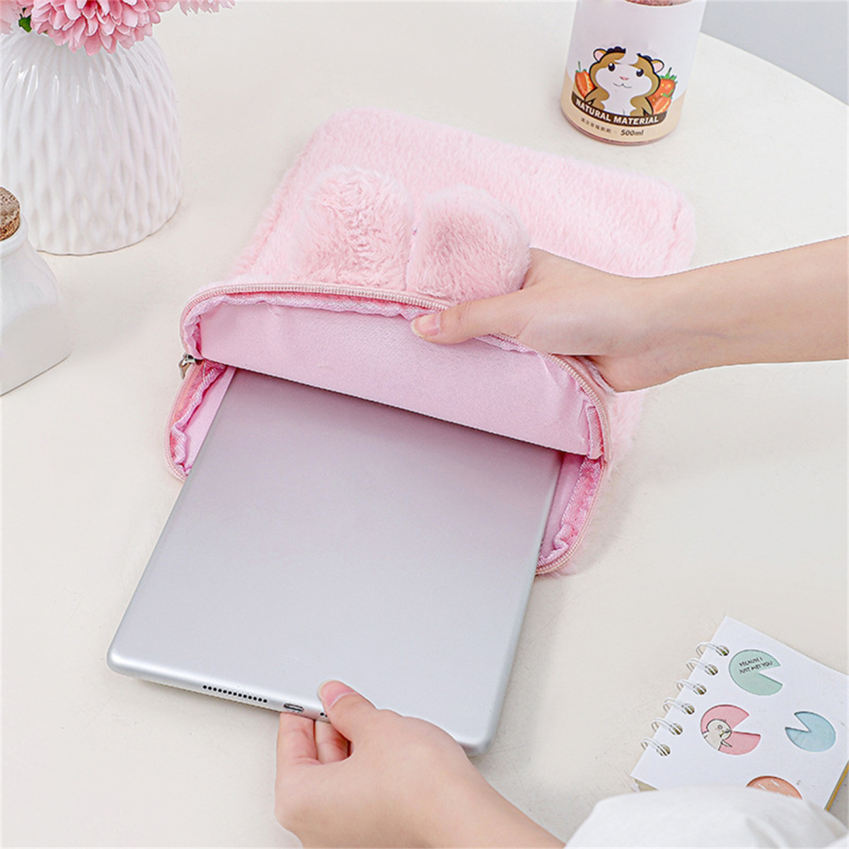 Eine Person legt ein Tablet in eine rosa, flauschige Hülle mit Hasenohren. Weitere Objekte sind eine Vase mit Blumen und ein Getränk.