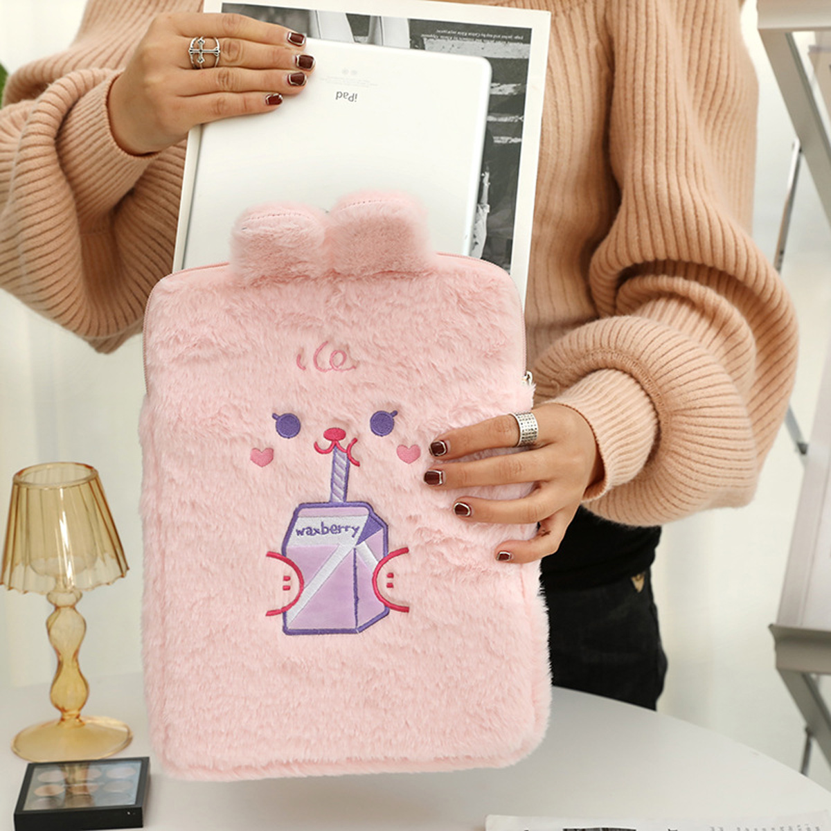 Person hält eine rosa, flauschige Hülle mit Cartoon-Gesicht und ein iPad. Die Hülle hat ein Saftbox-Design. Die Frau trägt einen Pullover und Ringe.