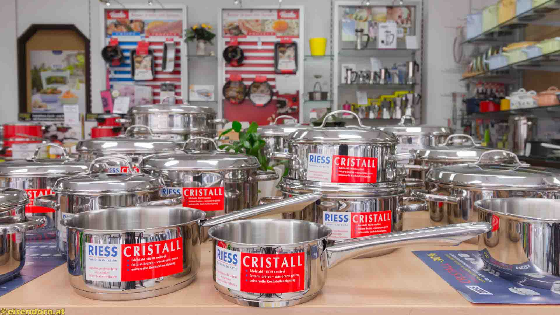 Eine Ladenausstellung zeigt zahlreiche Edelstahl-Töpfe und -Pfannen mit roten Etiketten. Regale im Hintergrund zeigen weiteres Kochgeschirr.