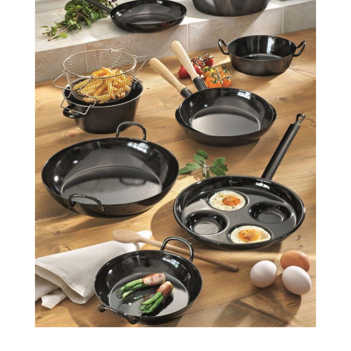 Eine Sammlung schwarzer Pfannen auf einer Holzoberfläche. Einige enthalten Lebensmittel wie Eier, Pommes Frites und Spargel. Eier und Tomaten sind ebenfalls vorhanden.