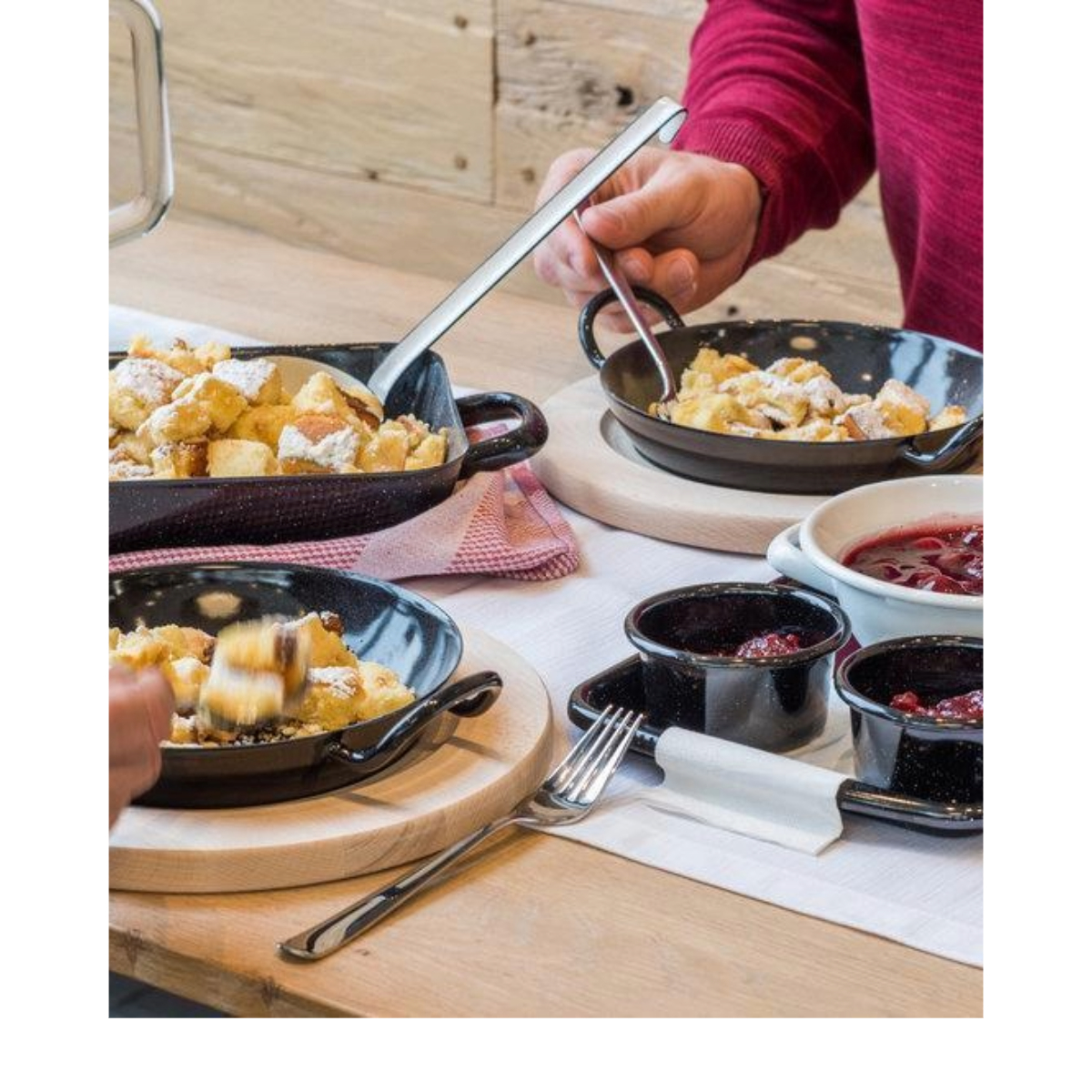 Ein Tisch mit mehreren Pfannen mit Essen und einer Person, die serviert. Es gibt kleine Schalen mit Soße, eine Gabel und eine Serviette.