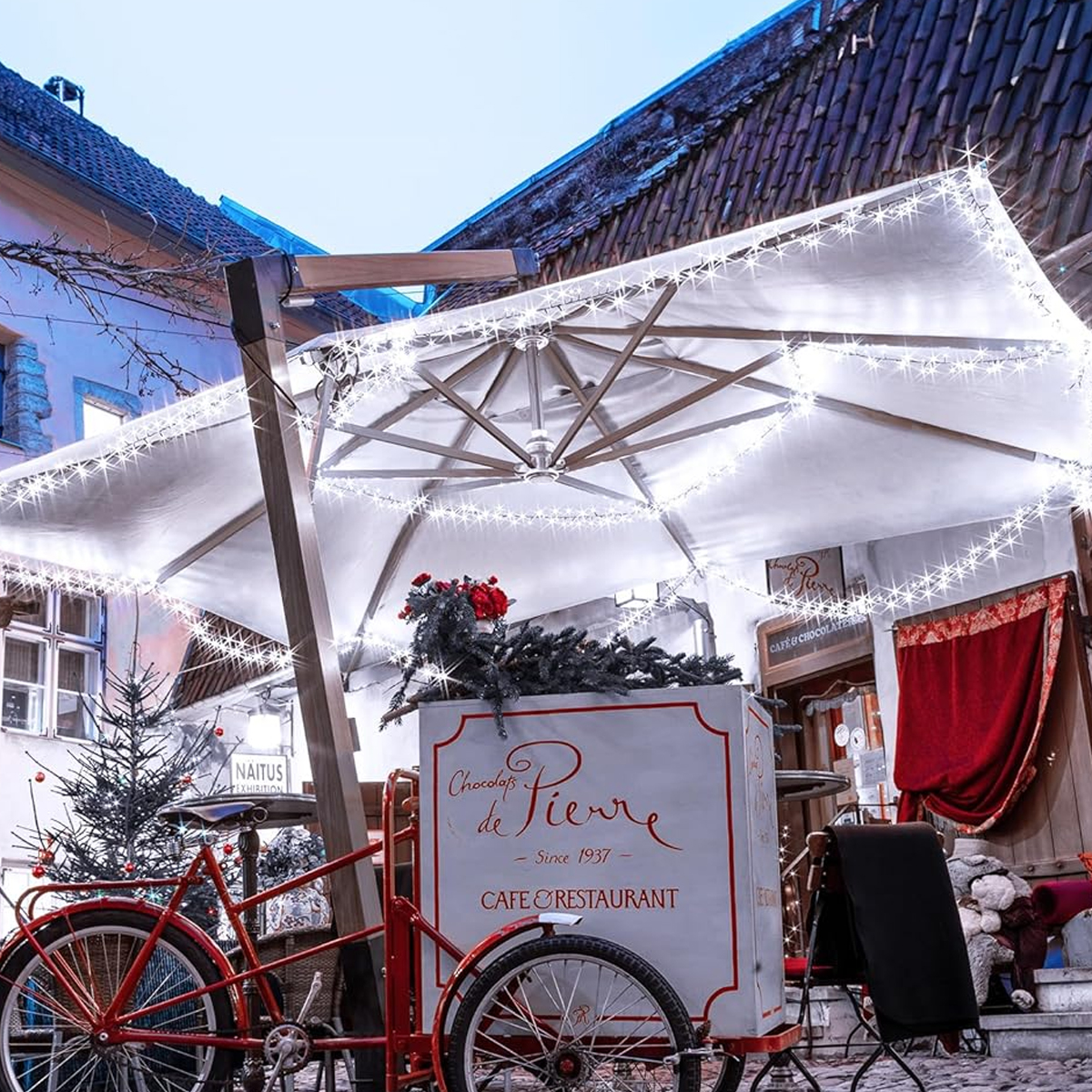 Ein roter Fahrradwagen mit Schirm, dekoriert mit Lichtern, steht vor einem Café. Ein Weihnachtsbaum ist sichtbar.