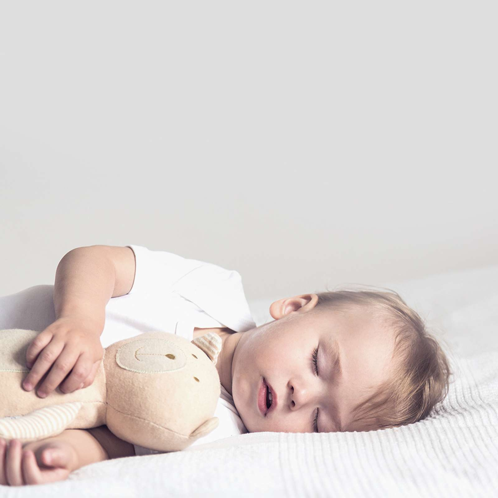 Ein schlafendes Baby liegt auf einem weißen Bett und hält ein beiges Stofftier. Das Baby trägt einen weißen Strampler.
