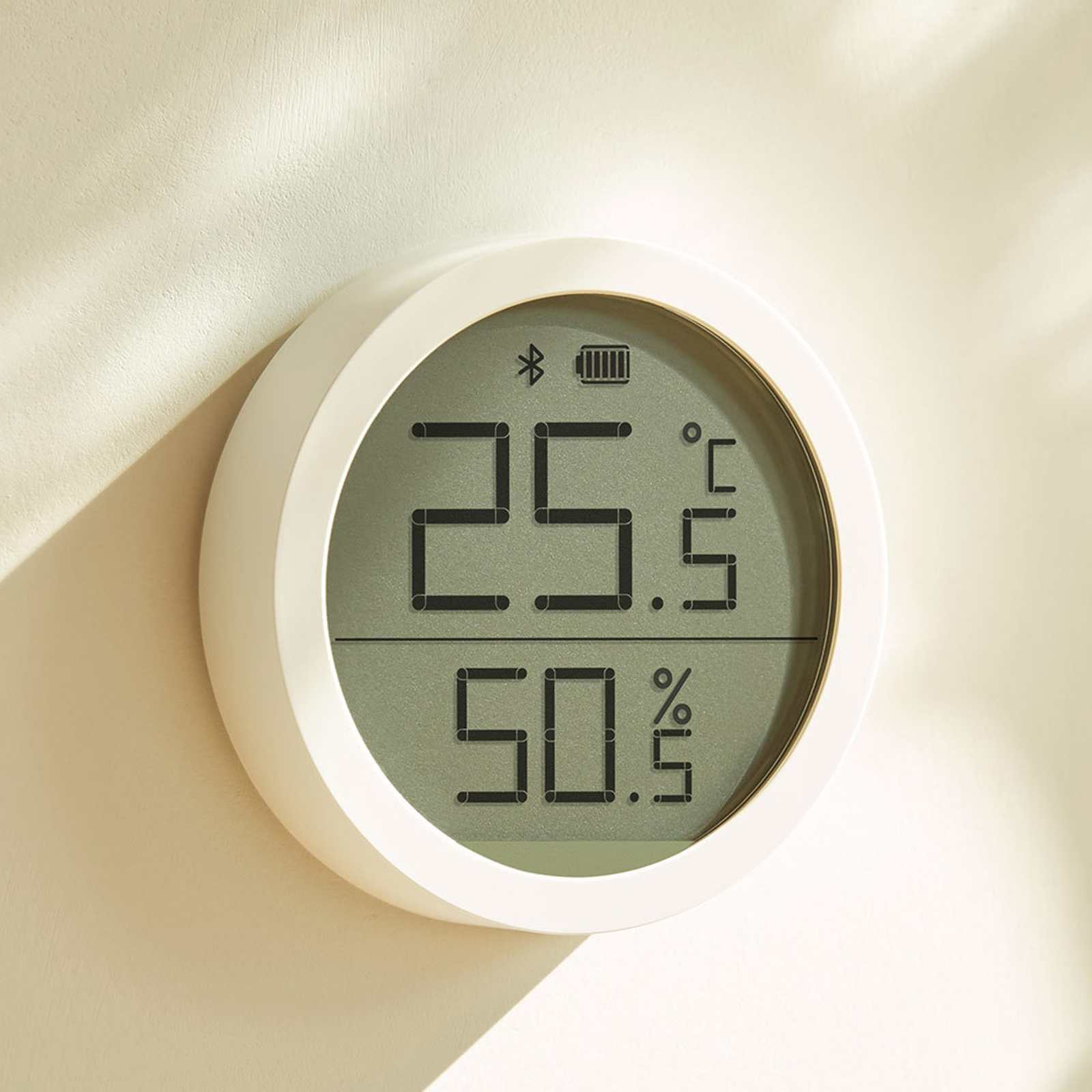 Ein weißes, rundes digitales Thermometer zeigt 25,5°C und 50,5% an. Es hat ein Bluetooth-Symbol und ein Akkusymbol. An einer beigen Wand montiert.