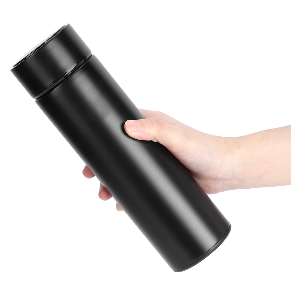 Eine Hand hält eine schwarze, isolierte Wasserflasche vor weißem Hintergrund. Die Flasche ist zylindrisch mit einem schwarzen Deckel.
