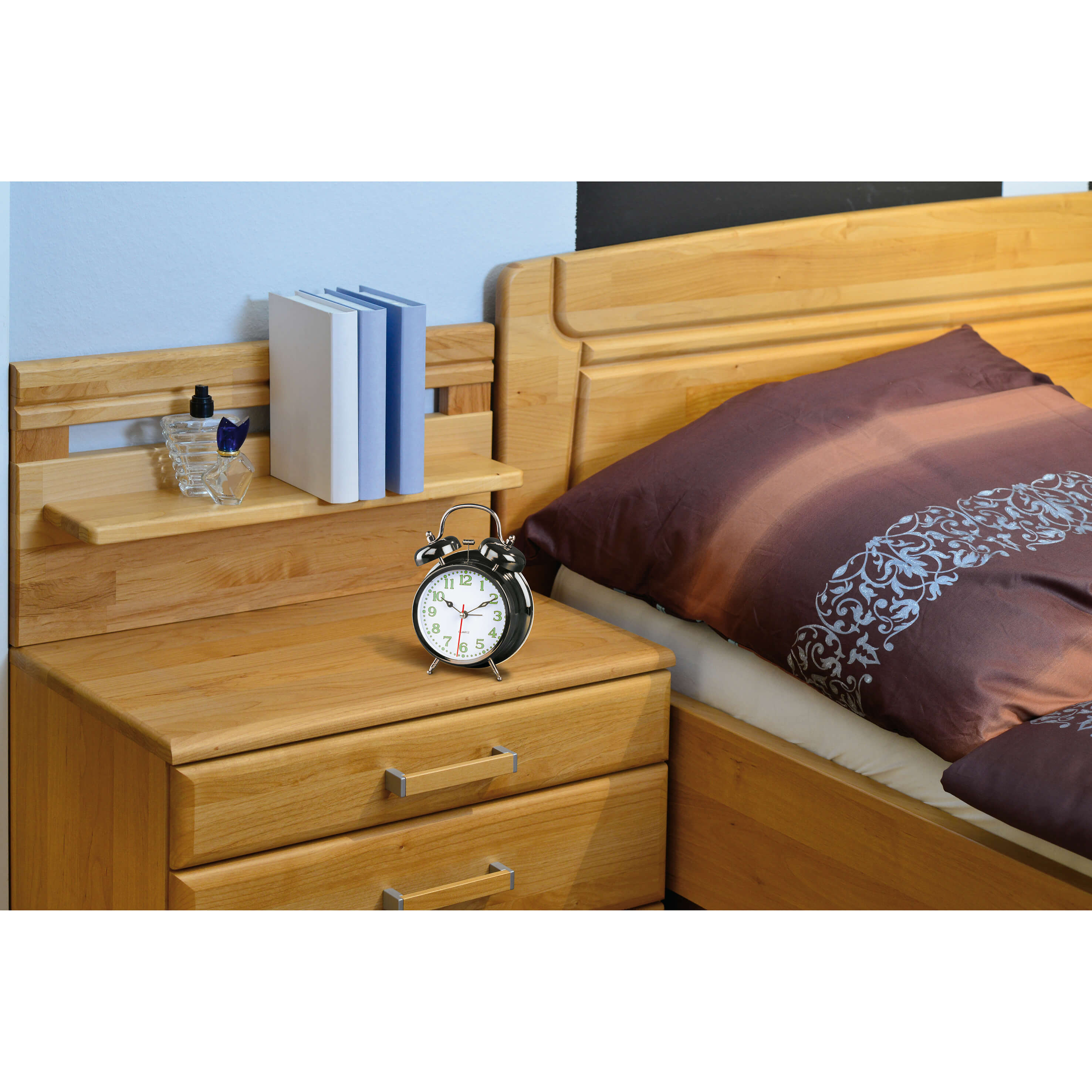 Ein Nachttisch aus Holz mit Wecker, Büchern und Parfüm. Im Hintergrund ein Bett mit brauner Bettwäsche.