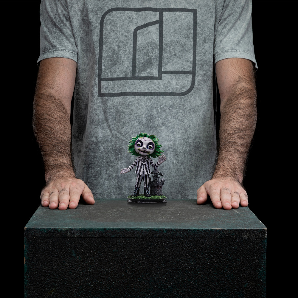 Una persona è in piedi dietro una statuina di Beetlejuice su una superficie verde. La statuina ha i capelli verdi e un completo a righe bianche e nere.
