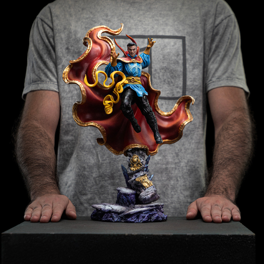 Eine Doctor Strange-Statue mit rotem Umhang und blauem Outfit steht auf einem Felsensockel. Eine Person mit grauem Hemd und behaarten Armen hält sie.