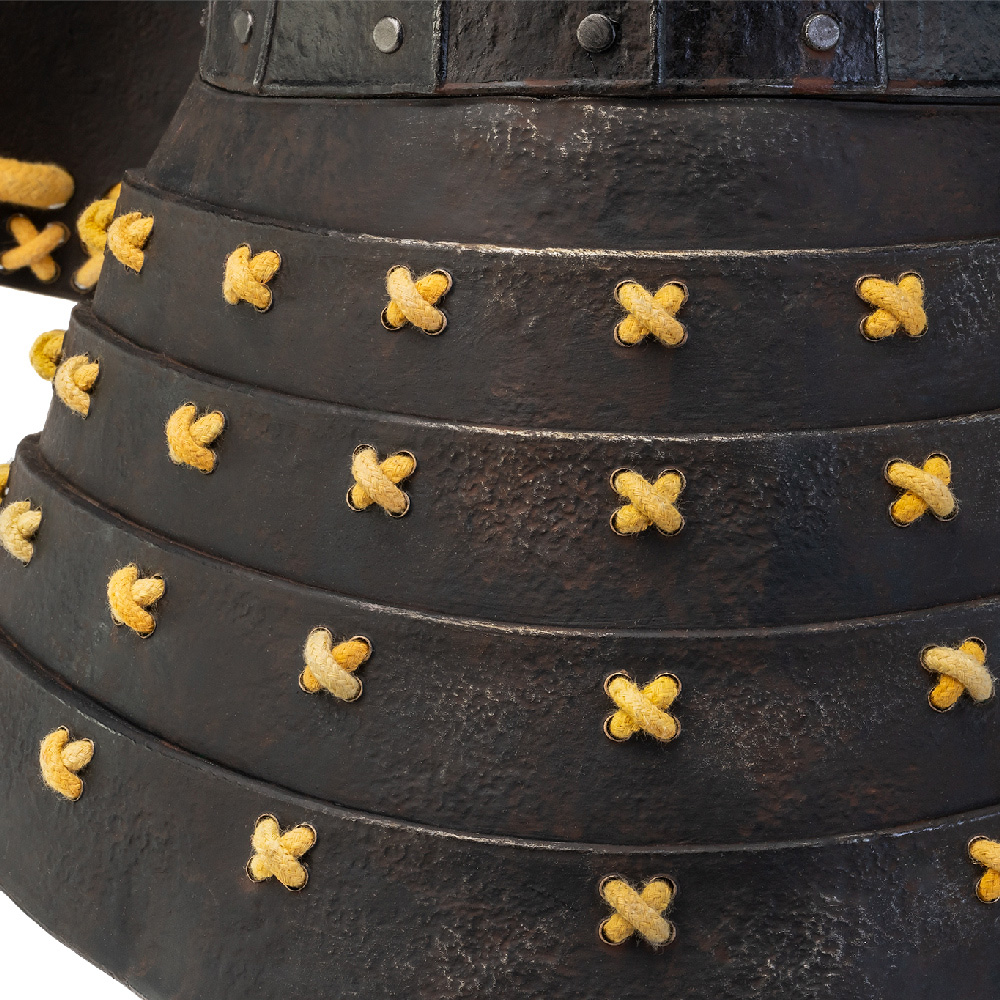 Nahaufnahme eines metallisch-schwarzen Samurai-Rüstungshelms mit gelben Seildetails und Nieten. Die Rüstung hat geschichtete Platten.