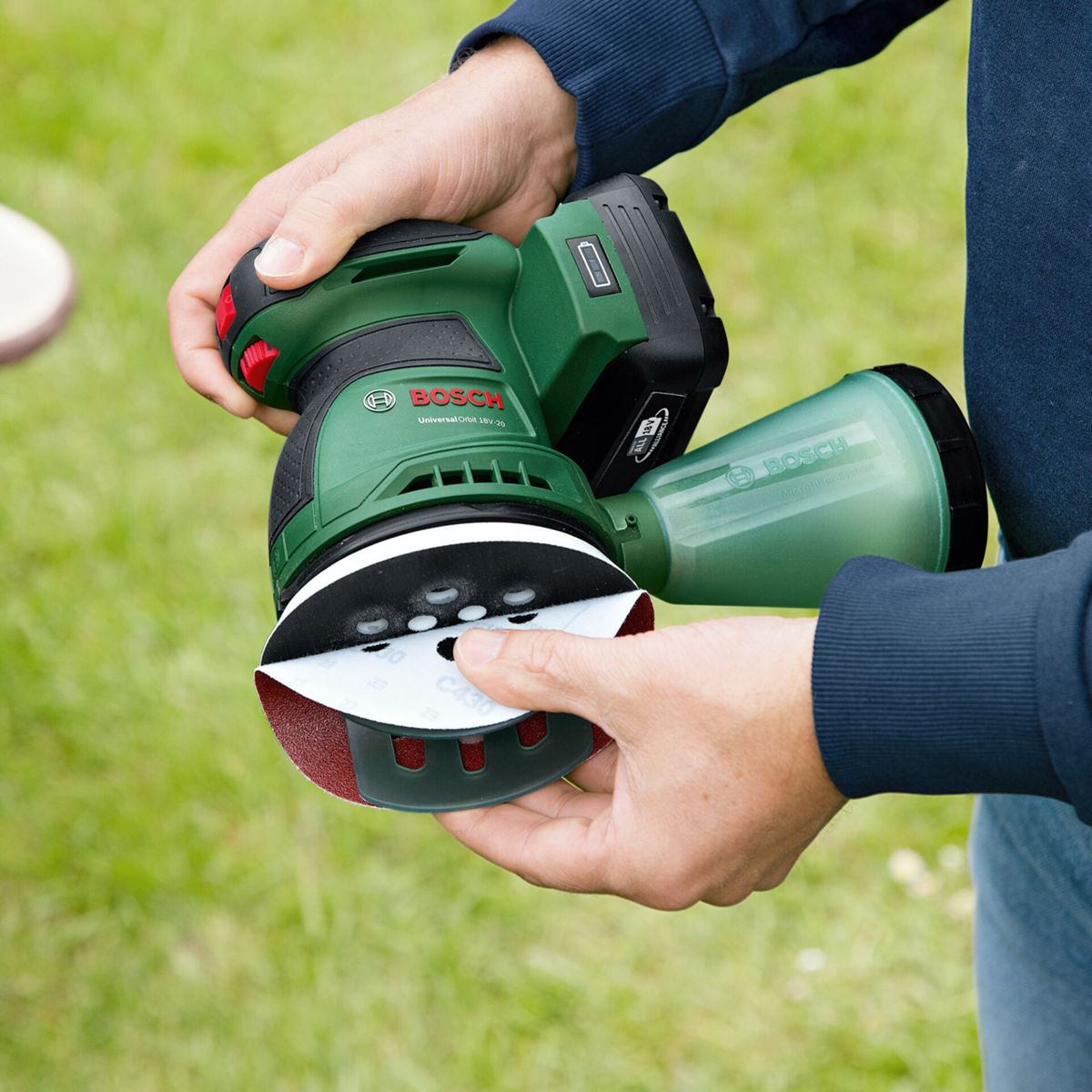 Une personne tient une ponceuse Bosch verte et noire avec un disque de ponçage rouge et blanc. De l'herbe verte en arrière-plan.