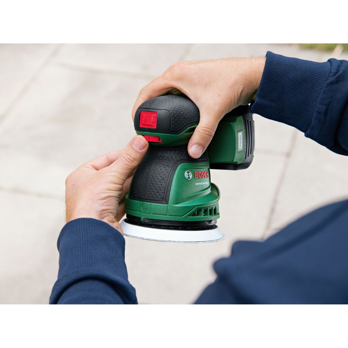 Une personne tient une ponceuse Bosch verte et noire. Les mains de la personne sont sur la ponceuse, et un interrupteur rouge est visible.