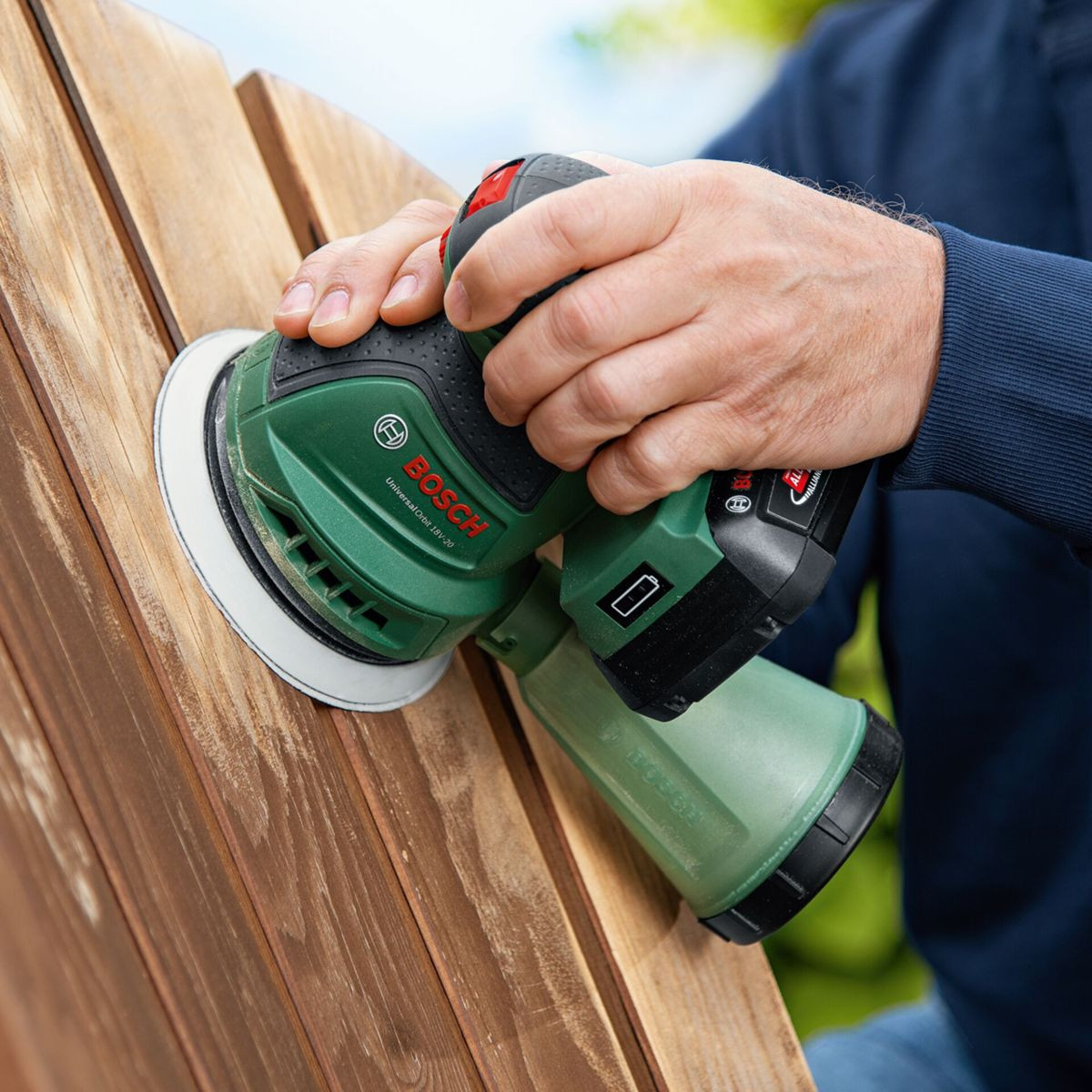 Une personne utilise une ponceuse Bosch verte sur une surface en bois. La ponceuse est tenue à deux mains. Le bois est brun.