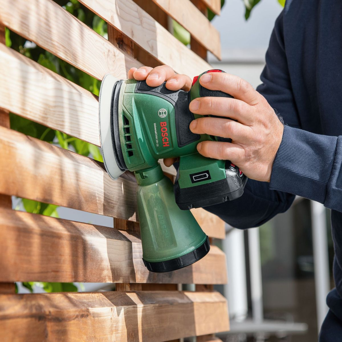 Une personne utilise une ponceuse Bosch verte sur une clôture en bois. La ponceuse est tenue à deux mains.