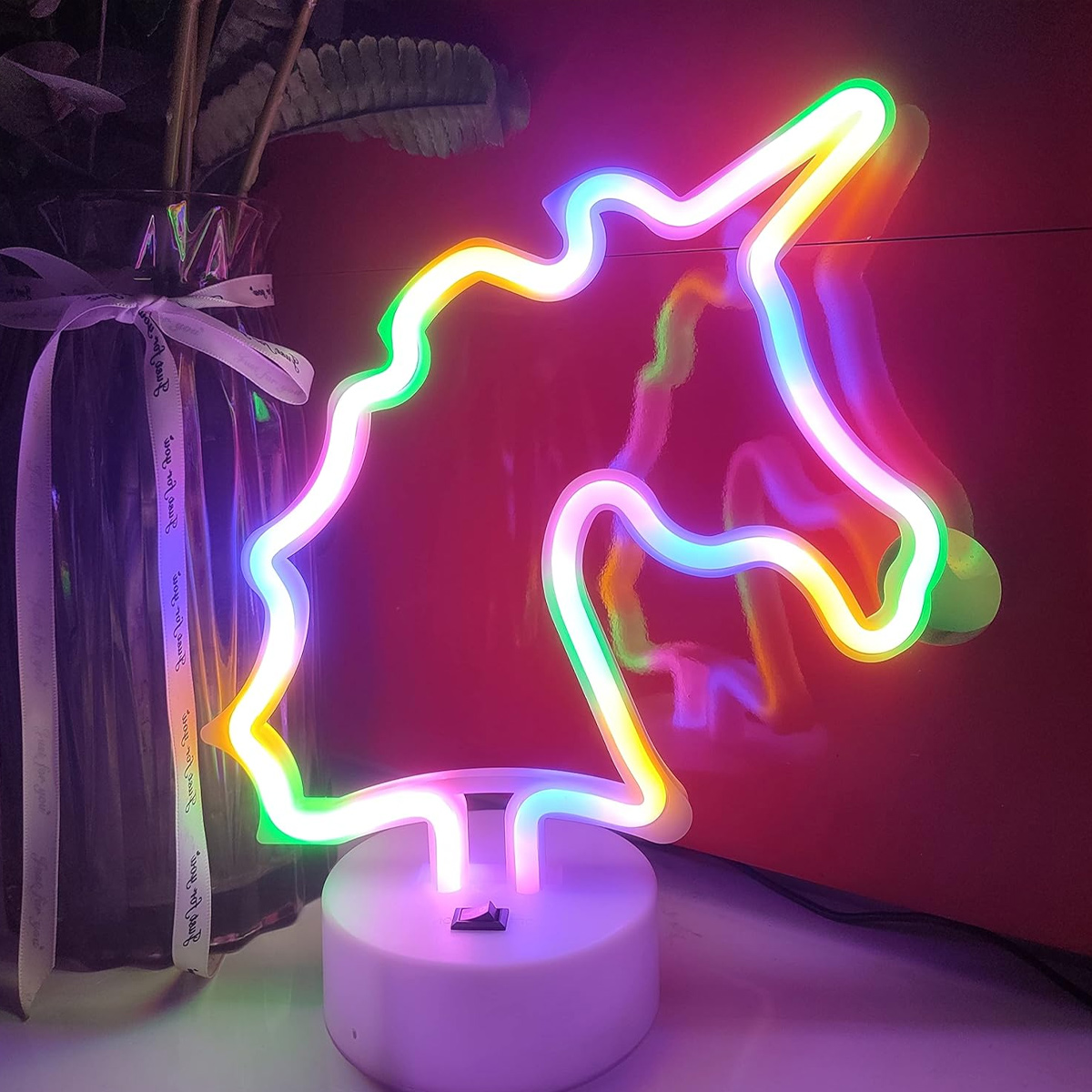 Ein Neon-Einhornlicht mit verschiedenen Farben auf weißer Basis. Links eine Vase mit Pflanzen. Der Hintergrund ist rot.
