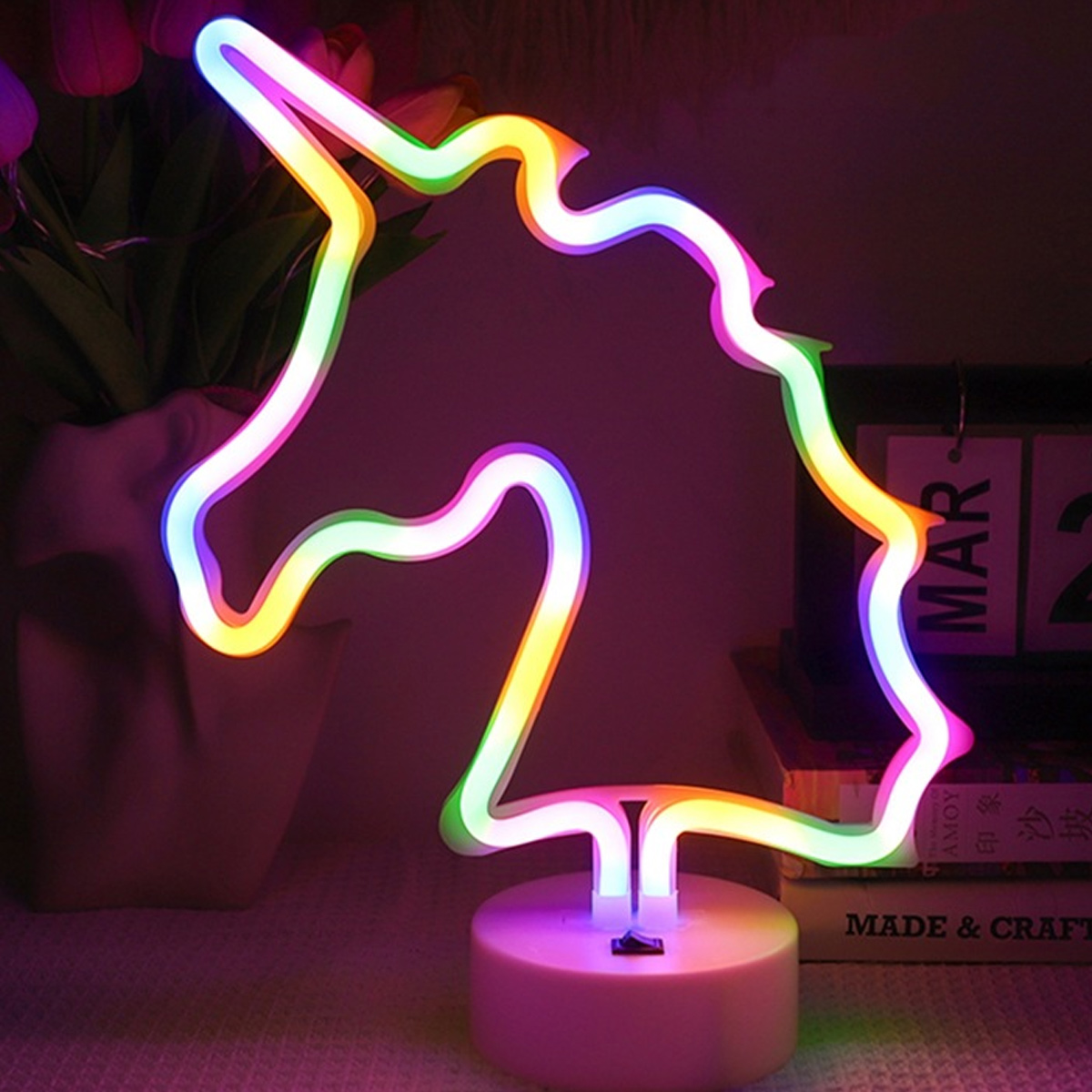 Eine Neon-Einhorn-Lampe. Sie ist mehrfarbig und steht auf einem weißen Sockel. Tulpen und Bücher sind im Hintergrund.