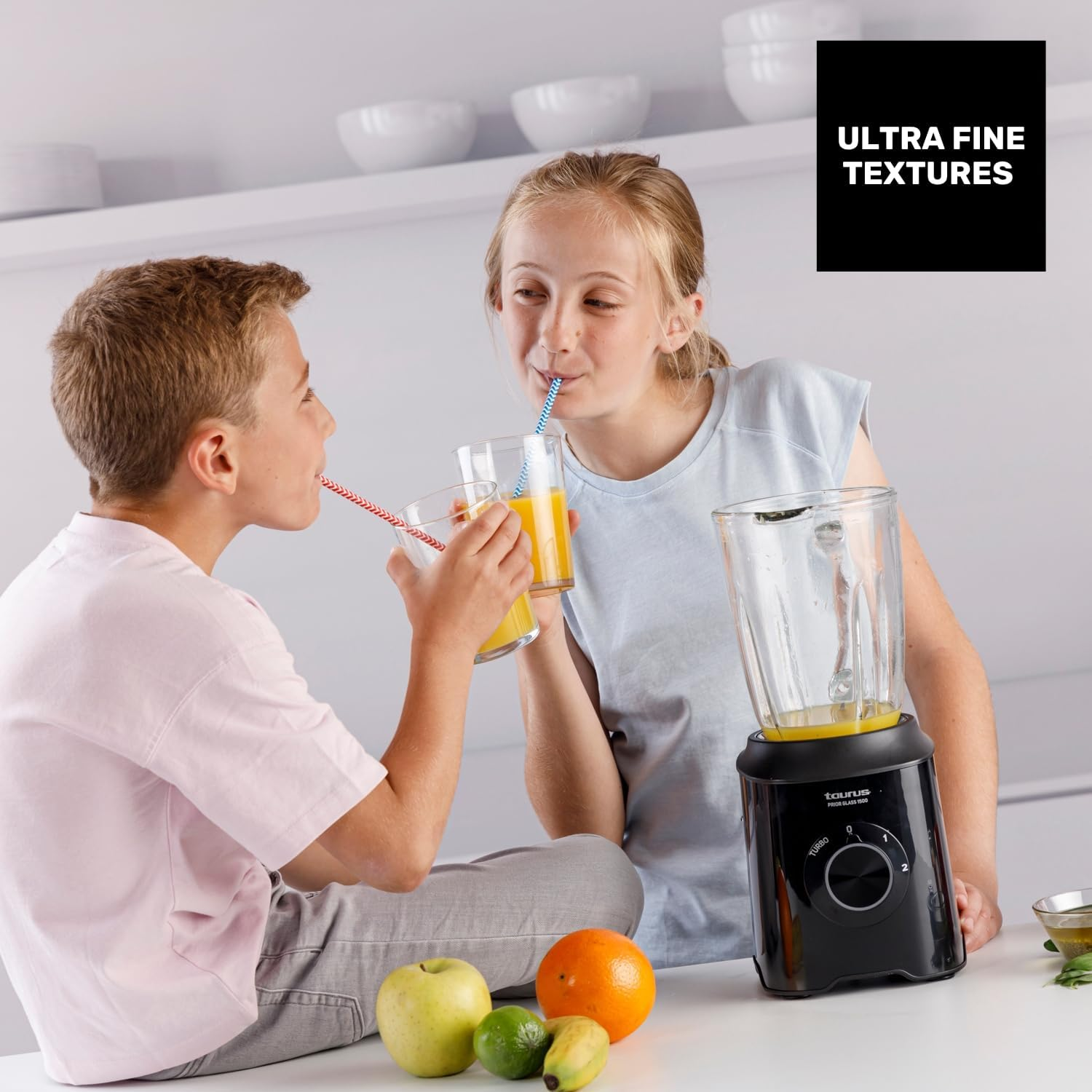 Dos niños bebiendo zumo con pajitas. Una licuadora está cerca. Frutas al frente. Armarios blancos al fondo.