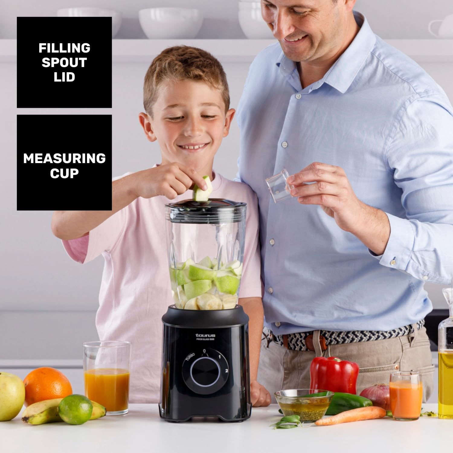 Un hombre y un niño preparan un batido. El niño pone una manzana en la licuadora. Frutas y verduras están en la mesa.