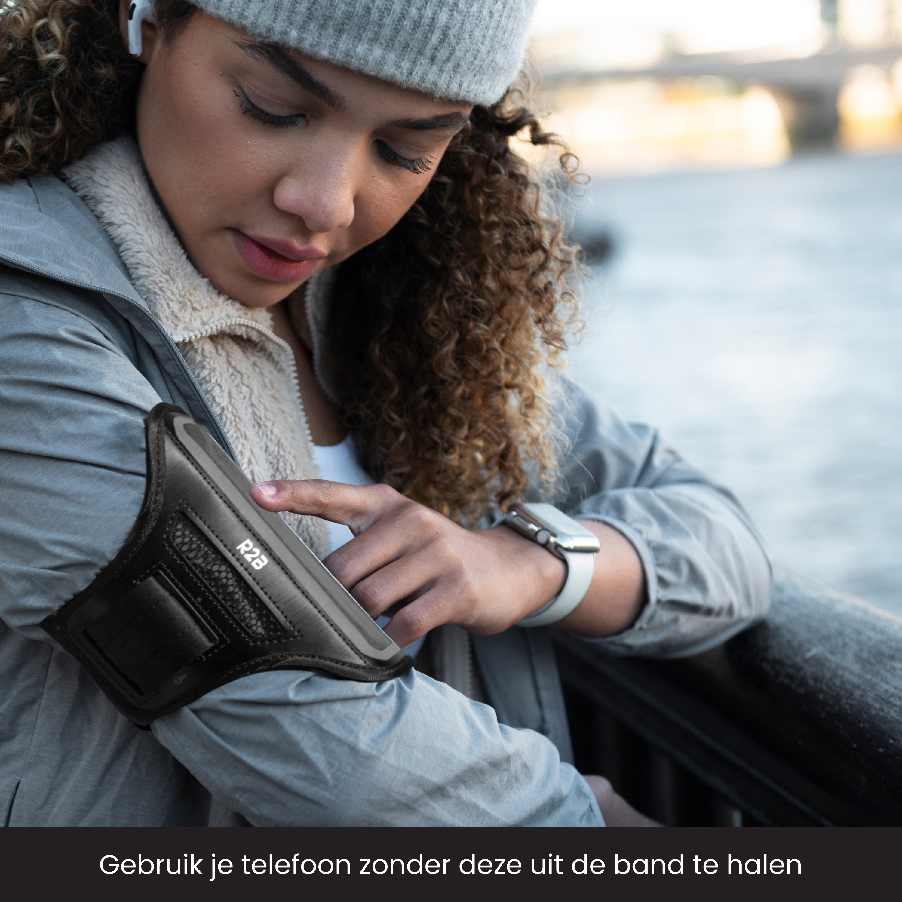 Frau benutzt Telefon in Armband, trägt Hut und Jacke. Im Hintergrund Fluss und Brücke.
