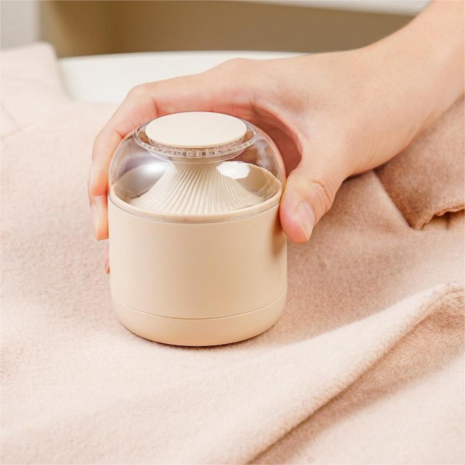 Hand hält einen beigefarbenen Stoffrasierer. Der Rasierer ist beige und aus klarem Kunststoff. Er liegt auf einer hellrosa Decke.