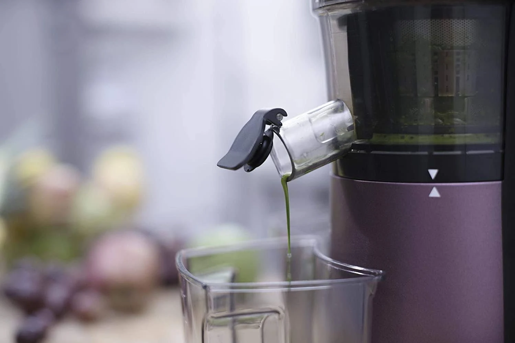 Una exprimidora dispensa jugo verde en un recipiente transparente. La exprimidora es morada y negra. Frutas borrosas en el fondo.