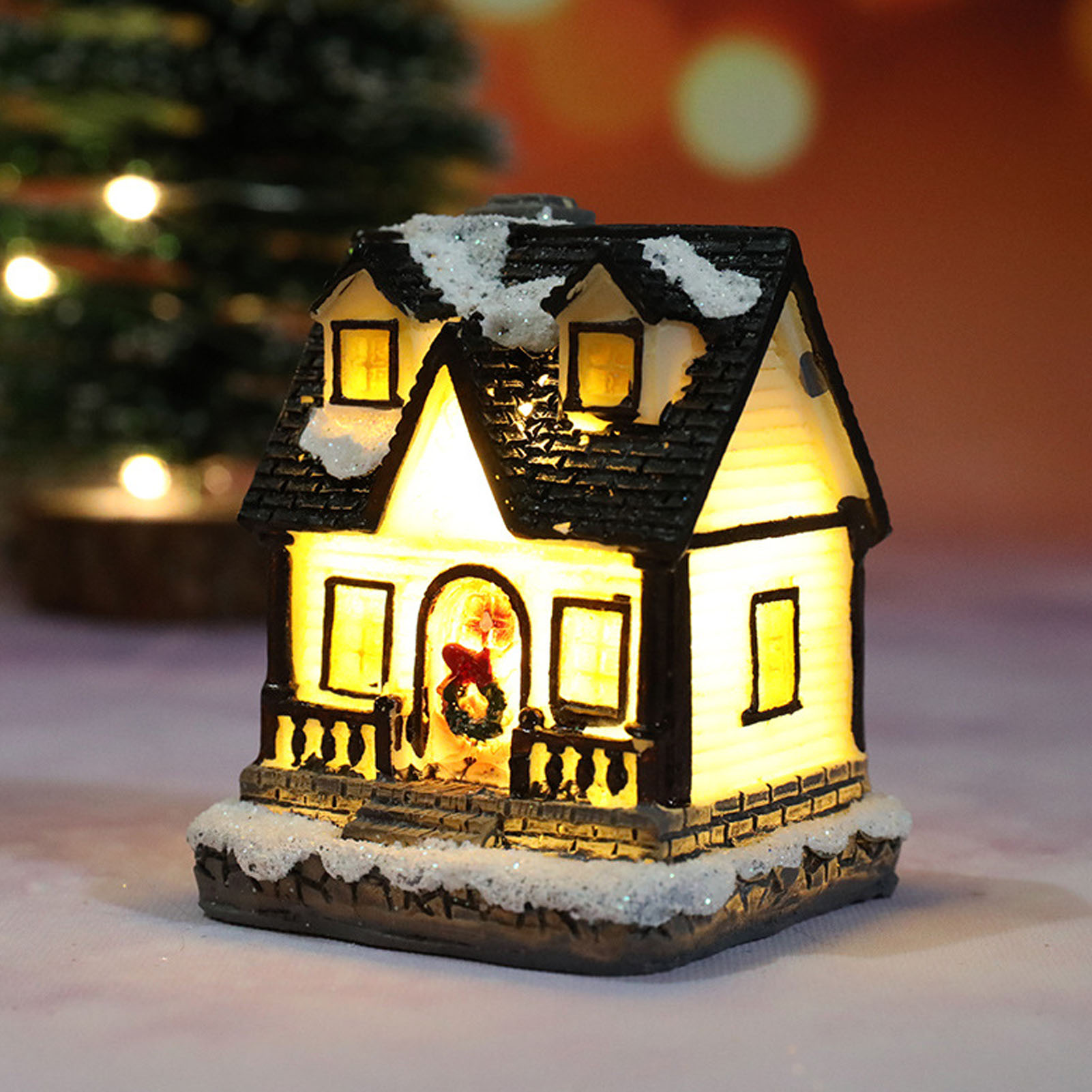 Ein beleuchtetes Miniatur-Weihnachtshaus mit Schnee, einem Kranz und einem Baum im Hintergrund.