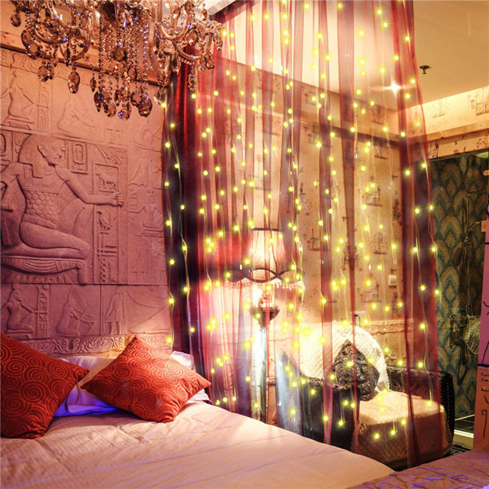 Schlafzimmer mit Bett, roten Kissen und transparenten Vorhängen mit Lichtern. Ein Kronleuchter hängt oben. Wand mit antiken Schnitzereien.