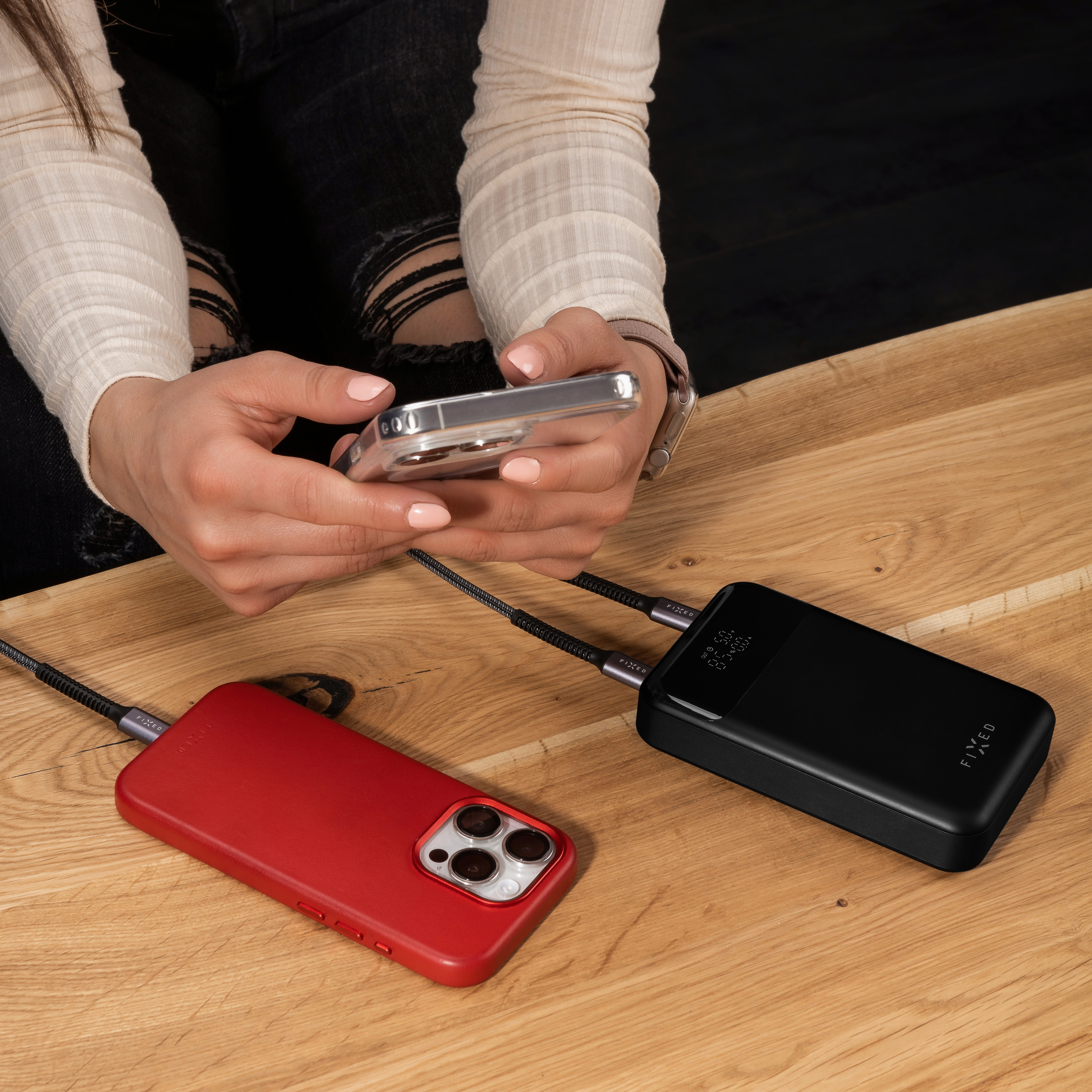 Eine Person benutzt ein Telefon und eine Powerbank. Eine rote Handyhülle liegt neben der schwarzen Powerbank auf einem Holztisch.