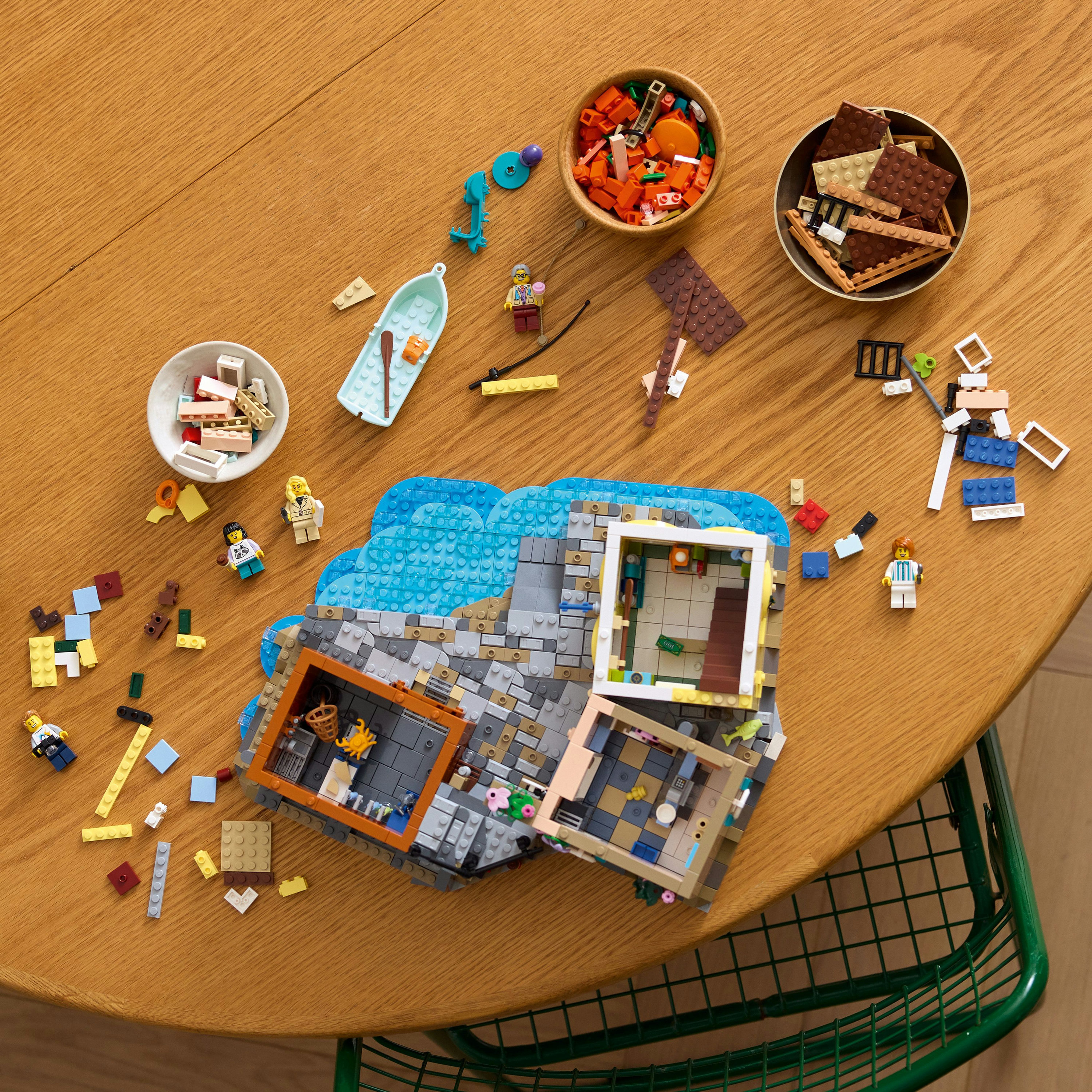 Zestaw Lego na drewnianym stole. Zawiera dom, łódź, figurki i elementy w miskach. Widoczne zielone krzesło.