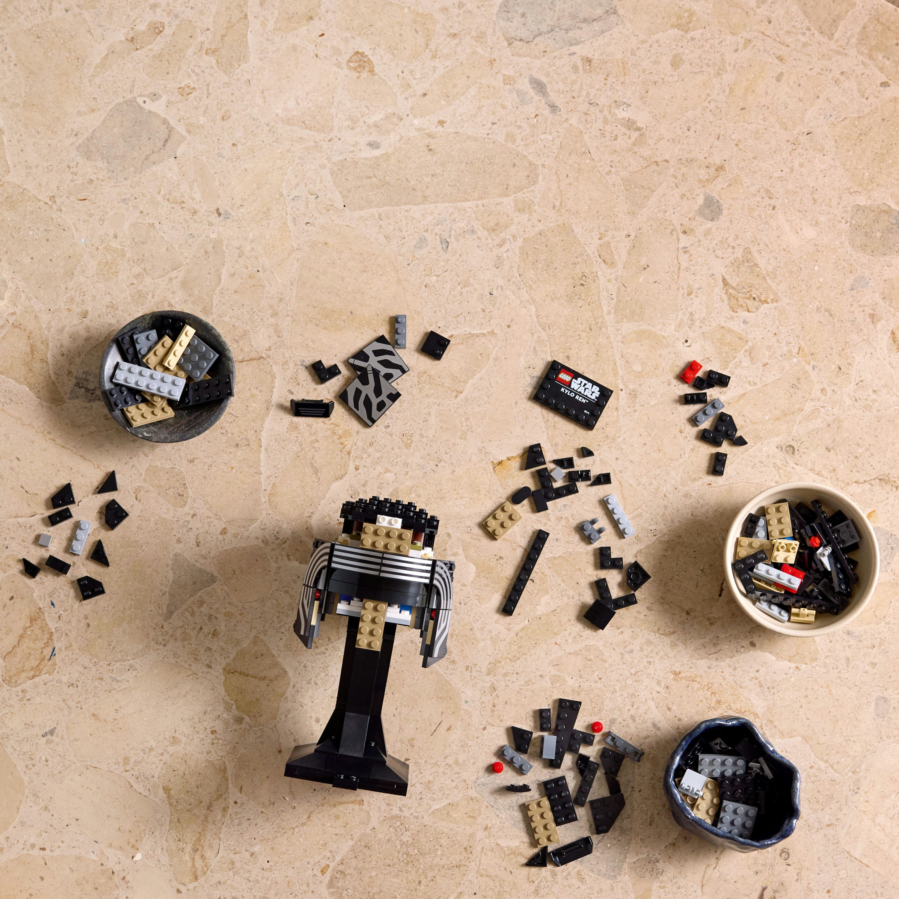 Rozłożona figurka Lego Kylo Ren na marmurze. Różne klocki Lego i małe miseczki rozrzucone wokół.