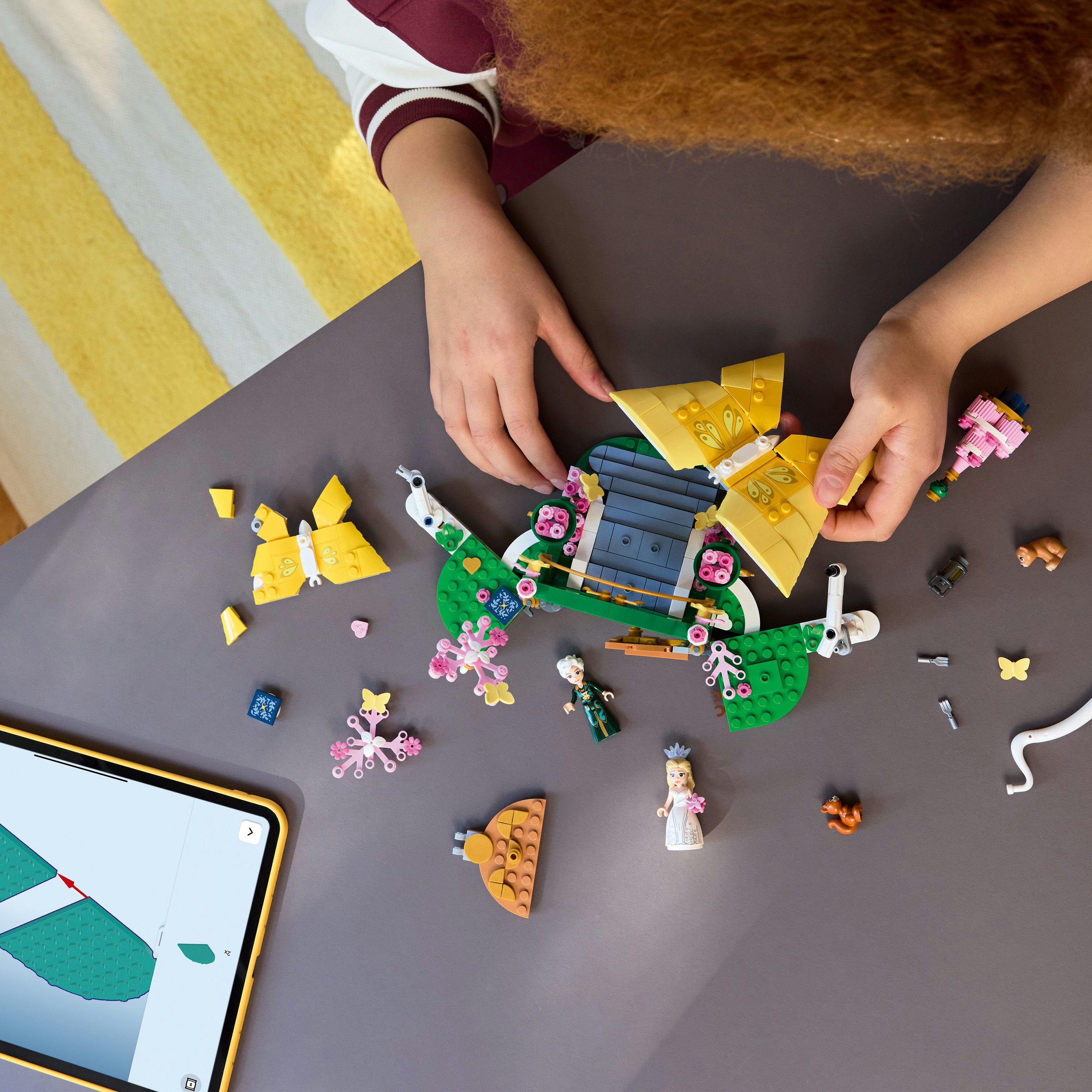 Una persona assembla un set Lego su un tavolo grigio. Il set presenta un ponte verde, ali di farfalla gialle e minifigure.
